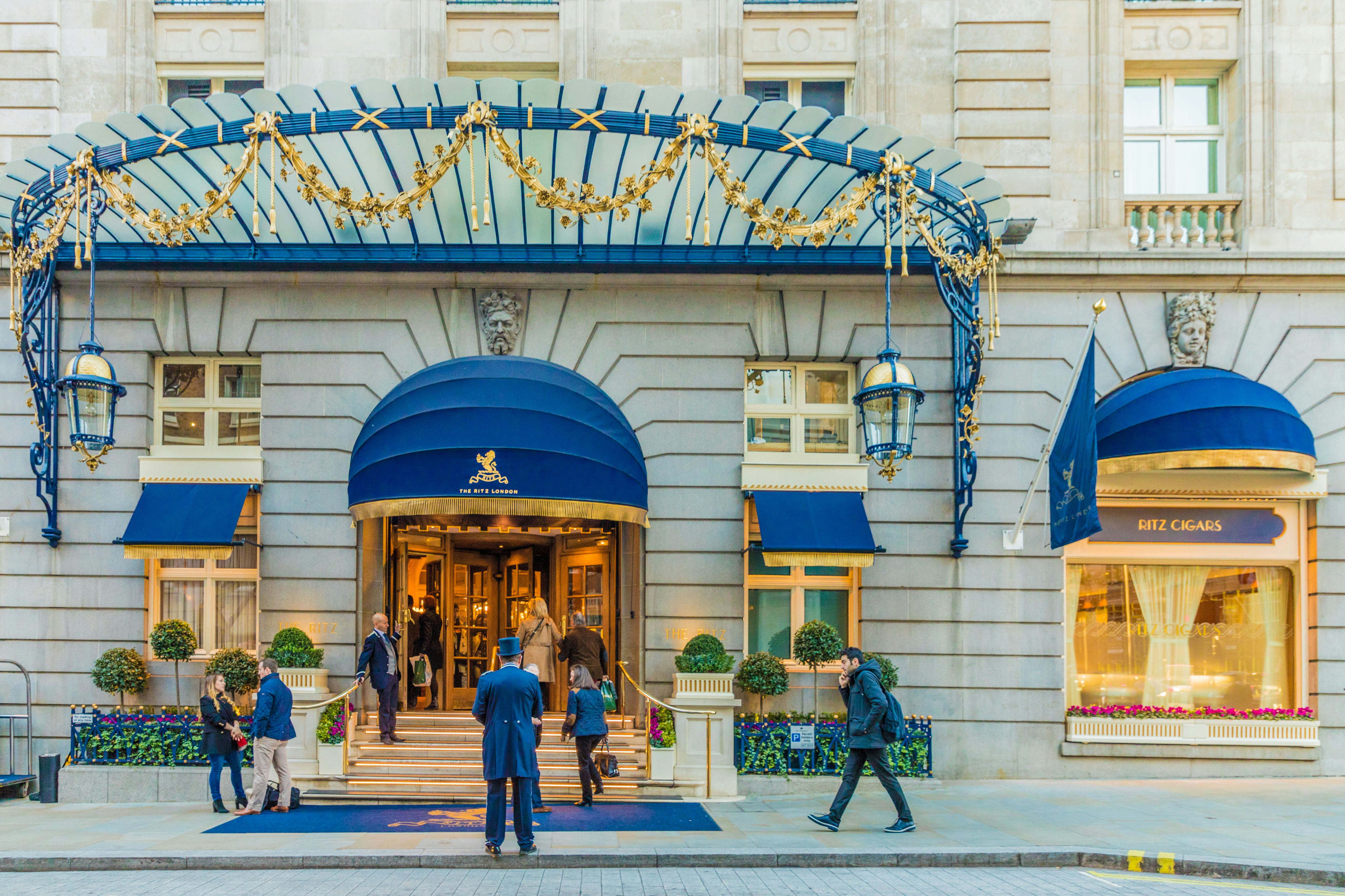 Stone facade of the hotel. There are blue and gold awnings, and staff waiting outside. The lights are on in the hotel.