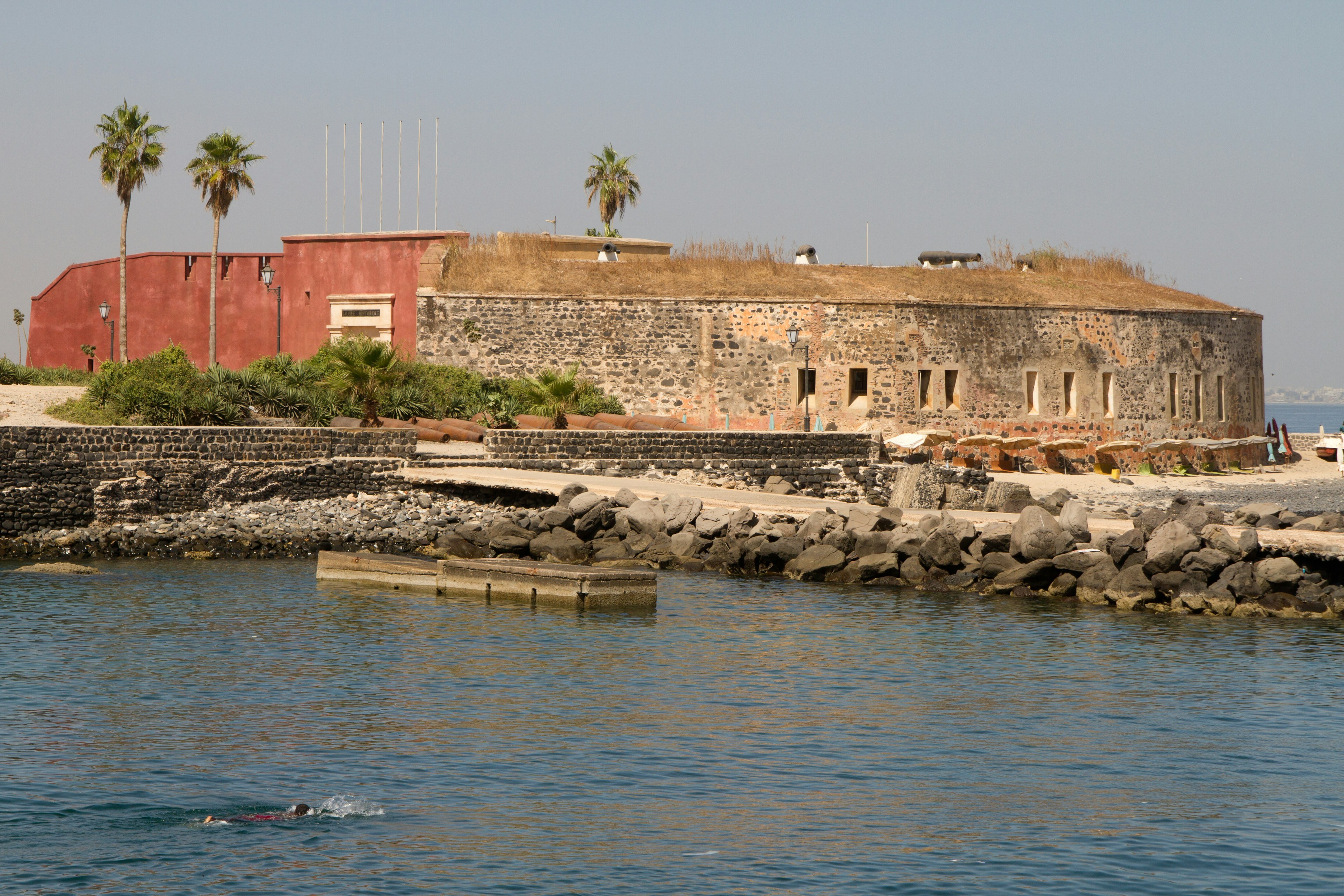 This image looks across the water to Île de Gorée and a circular stone fortress, with its small windows. To the left of the fortress is a pinkish section of the fortress, with palms in the background. A boat floats in the foreground.