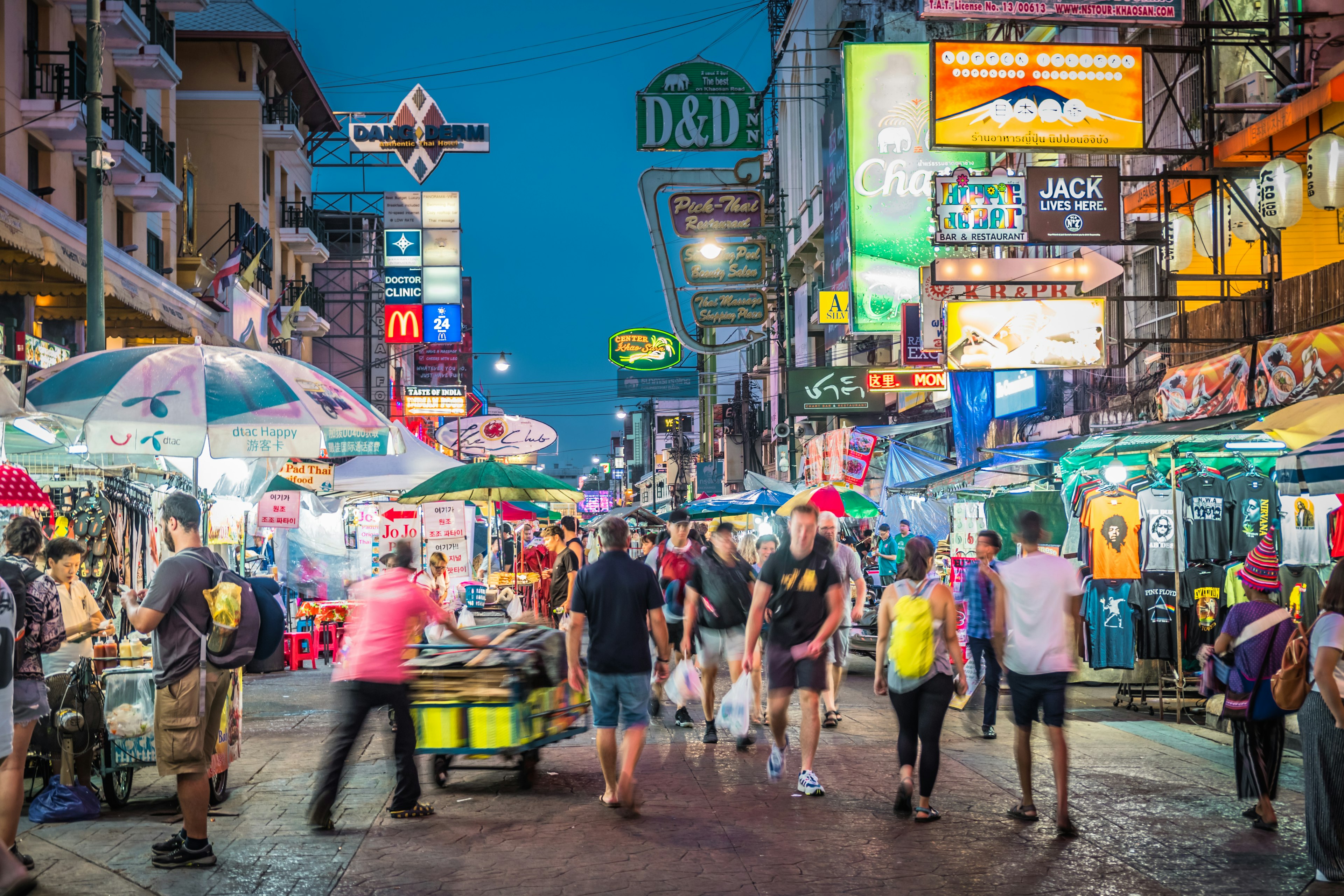 People make their way down a street lined with vendors selling food, clothes, drinks. Neon lights hang to the sides