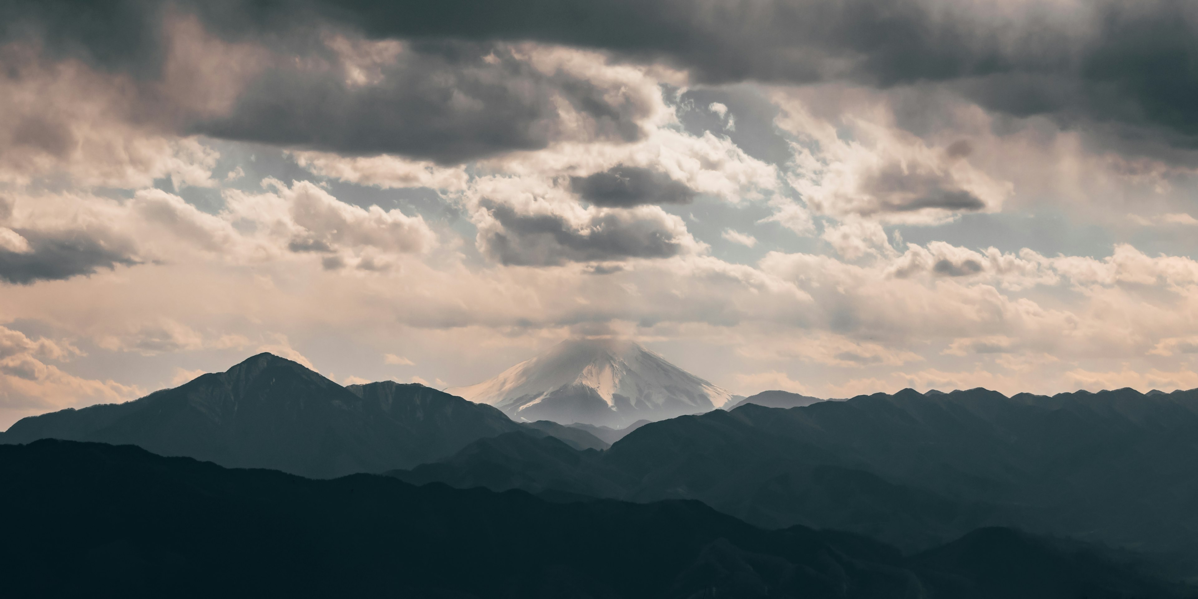 A distant view of Mt Fuji from Mt Takao. The iconic mountain rises above the surrounding hills and its snow-capped peak is enveloped by wispy white cloud.