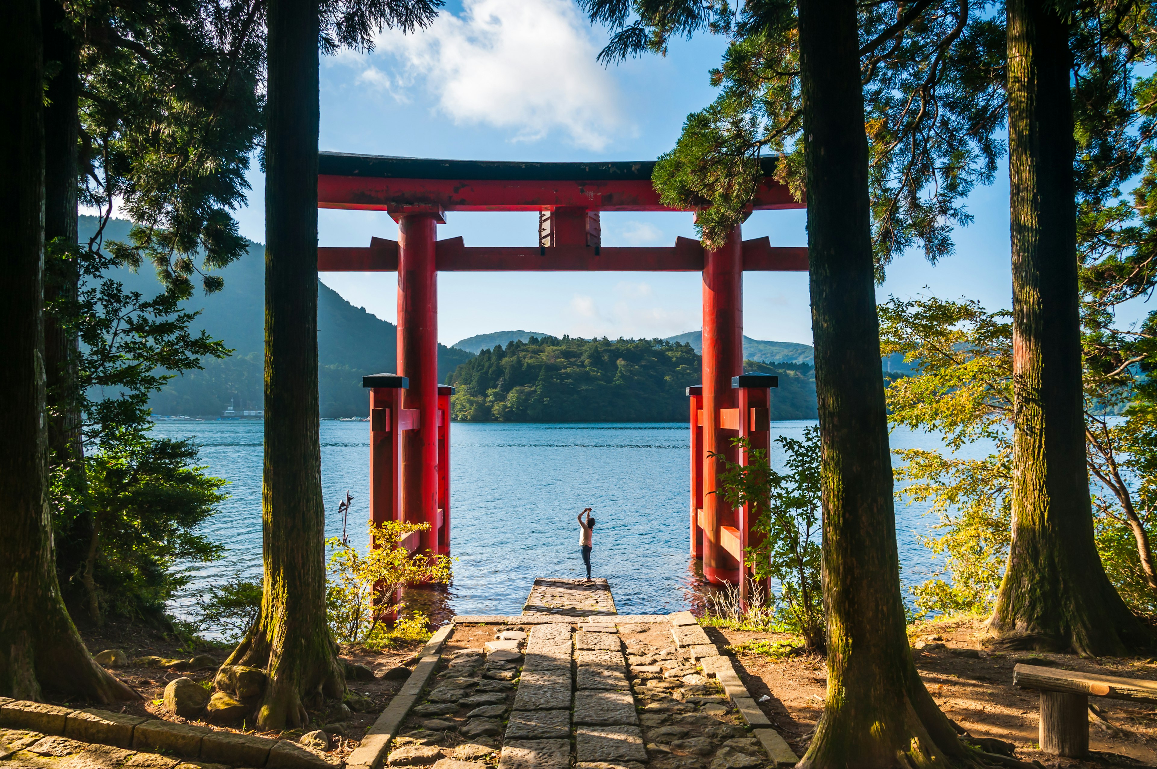 A woman takes photos with her phone of a Japanese red gateway leading to a picturesque lake