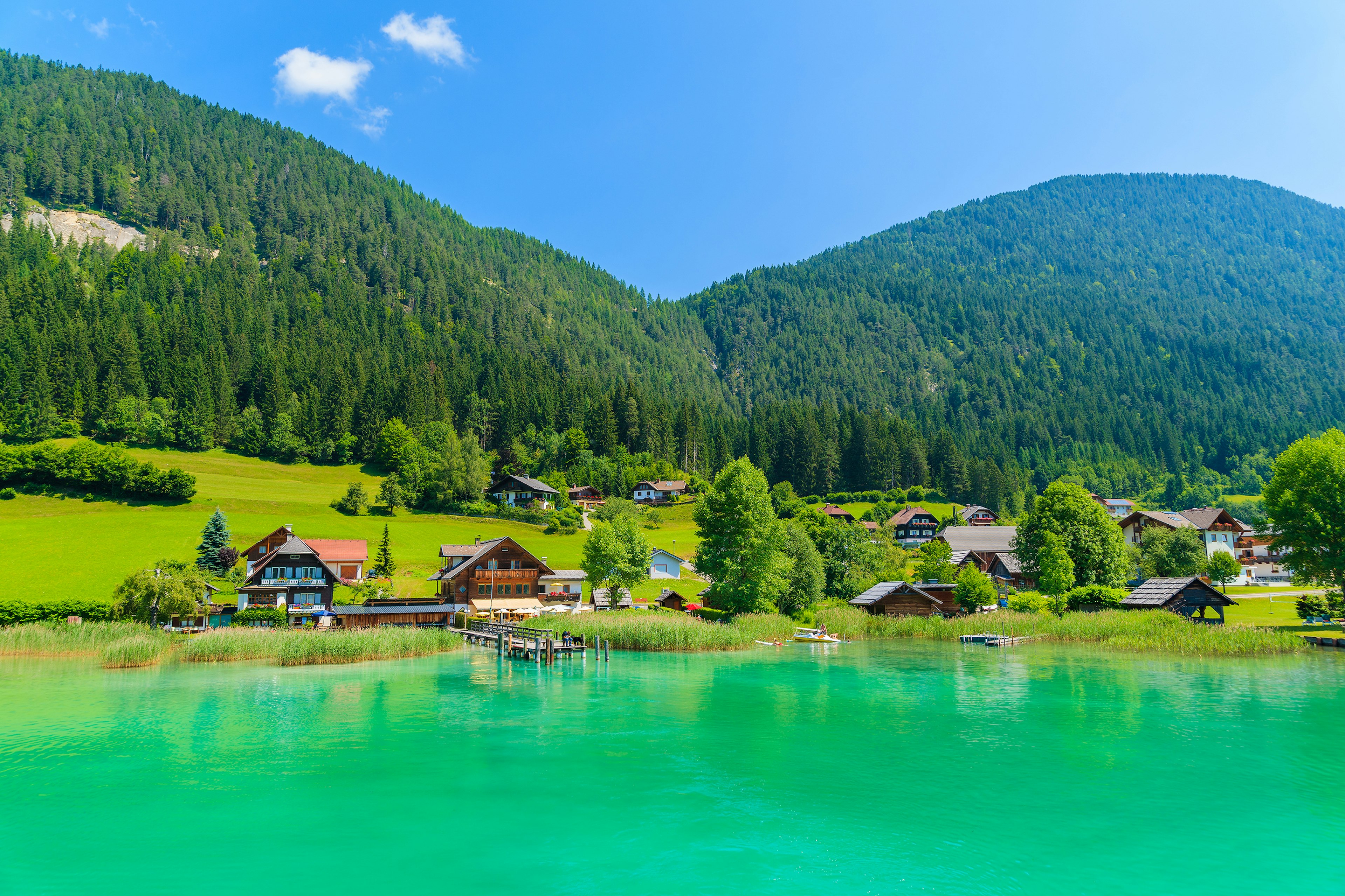 A bright green lake surrounded by hills covered in woodland. There are several chalet-style houses on the lakeside