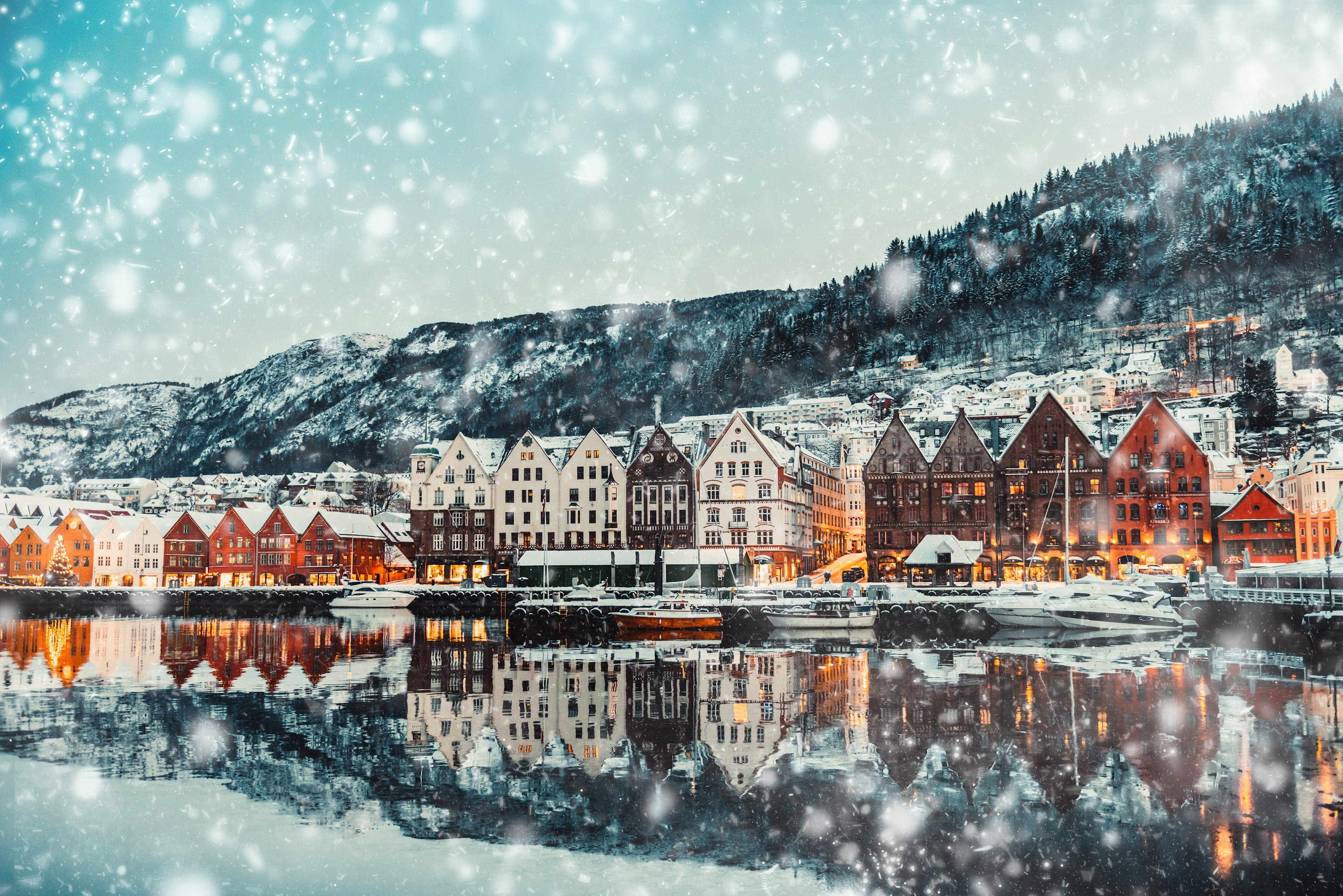 A row of colourful houses with pointed roofs stand alongside a dock. There is forest on the hills behind and the snow is falling heavily