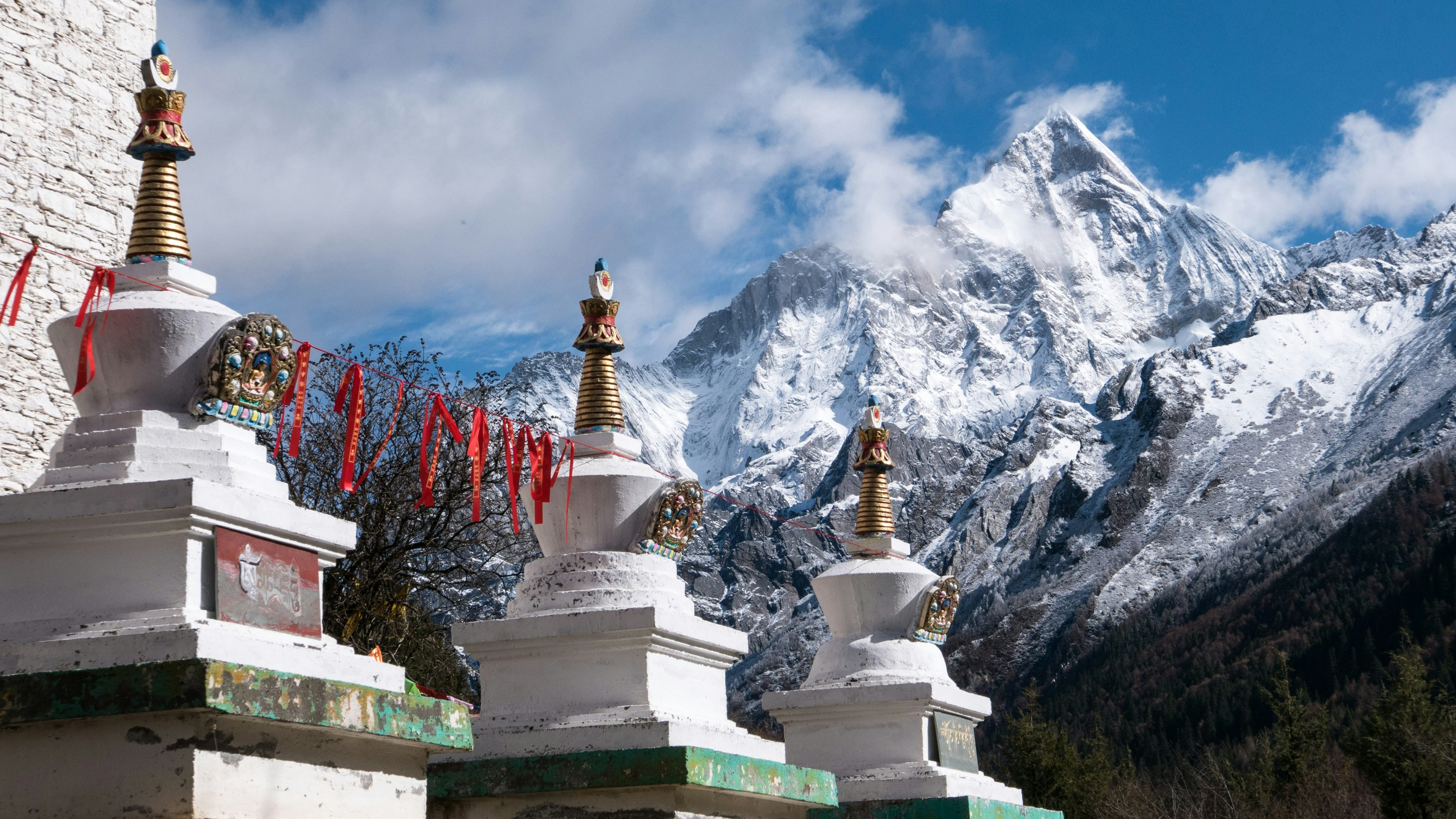 Snow covered Siguniang Mountain is seen in the background of Chinese temple, Lame Temple. Red streamers hang between three identical statues.