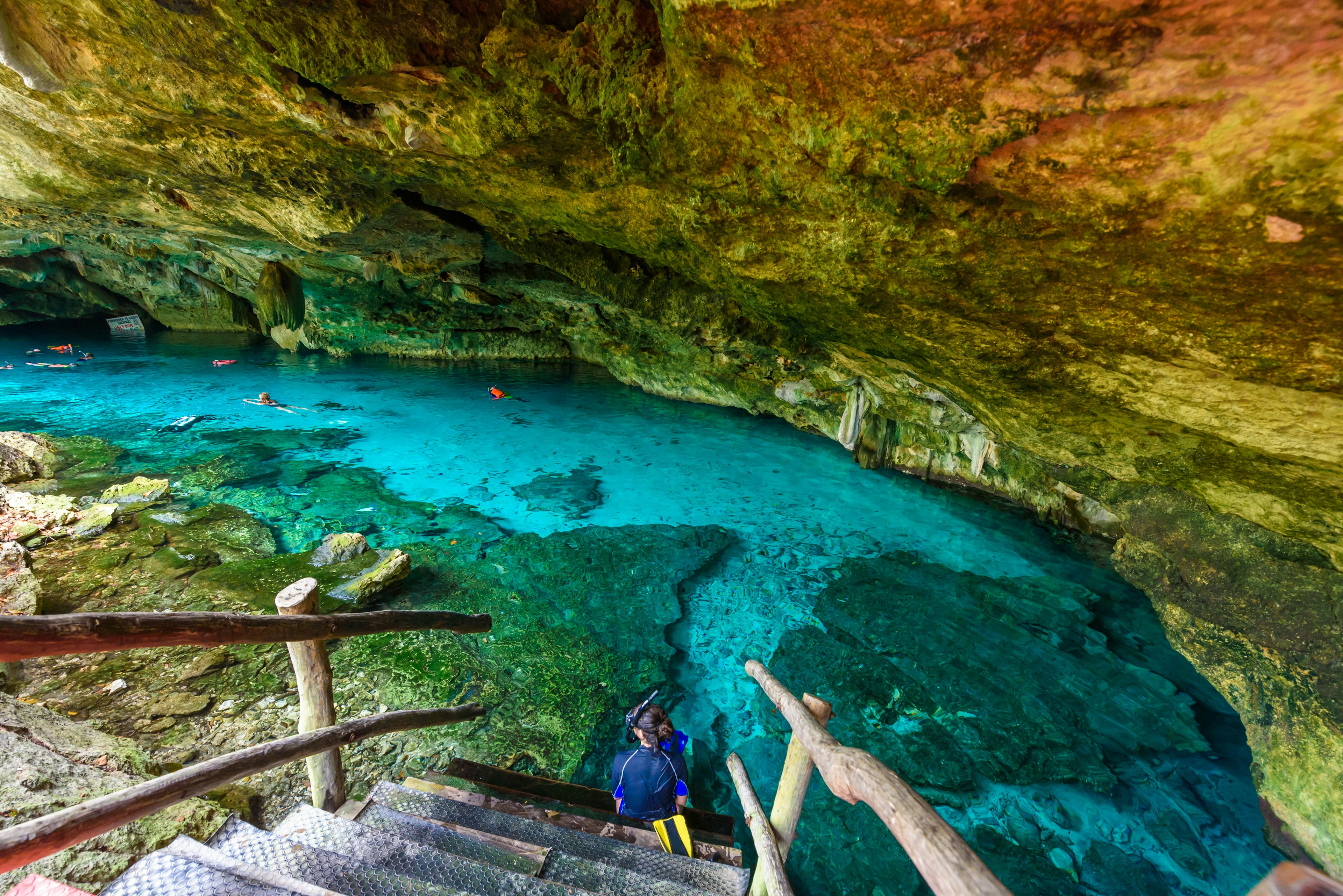 A snorkeler sits at the bottom of a wooden stairway leading into a blue pool in a cave