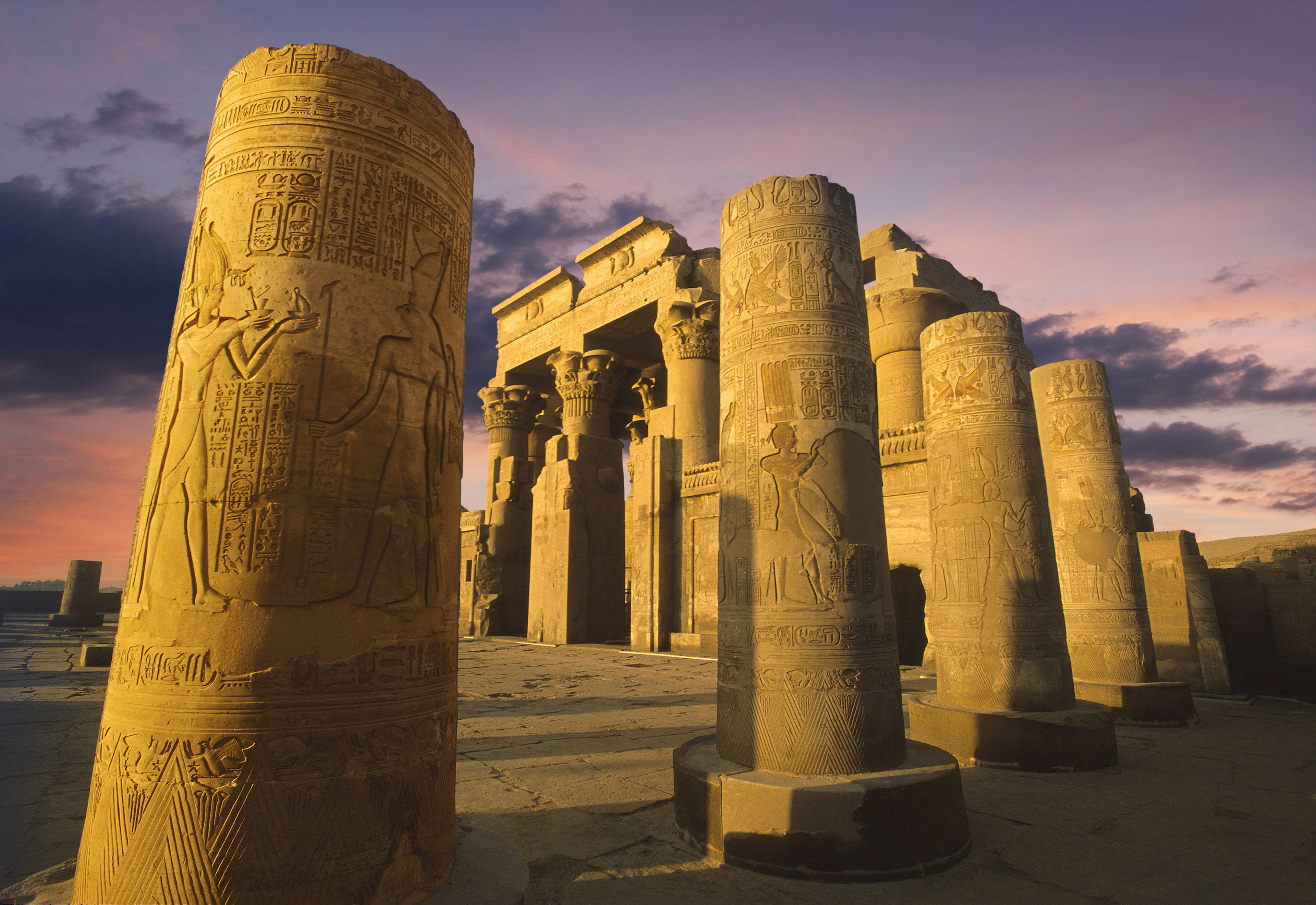 An Egyptian temple with large stone columns covered in etchings and hieroglyphics lit up in the evening as the sun sets