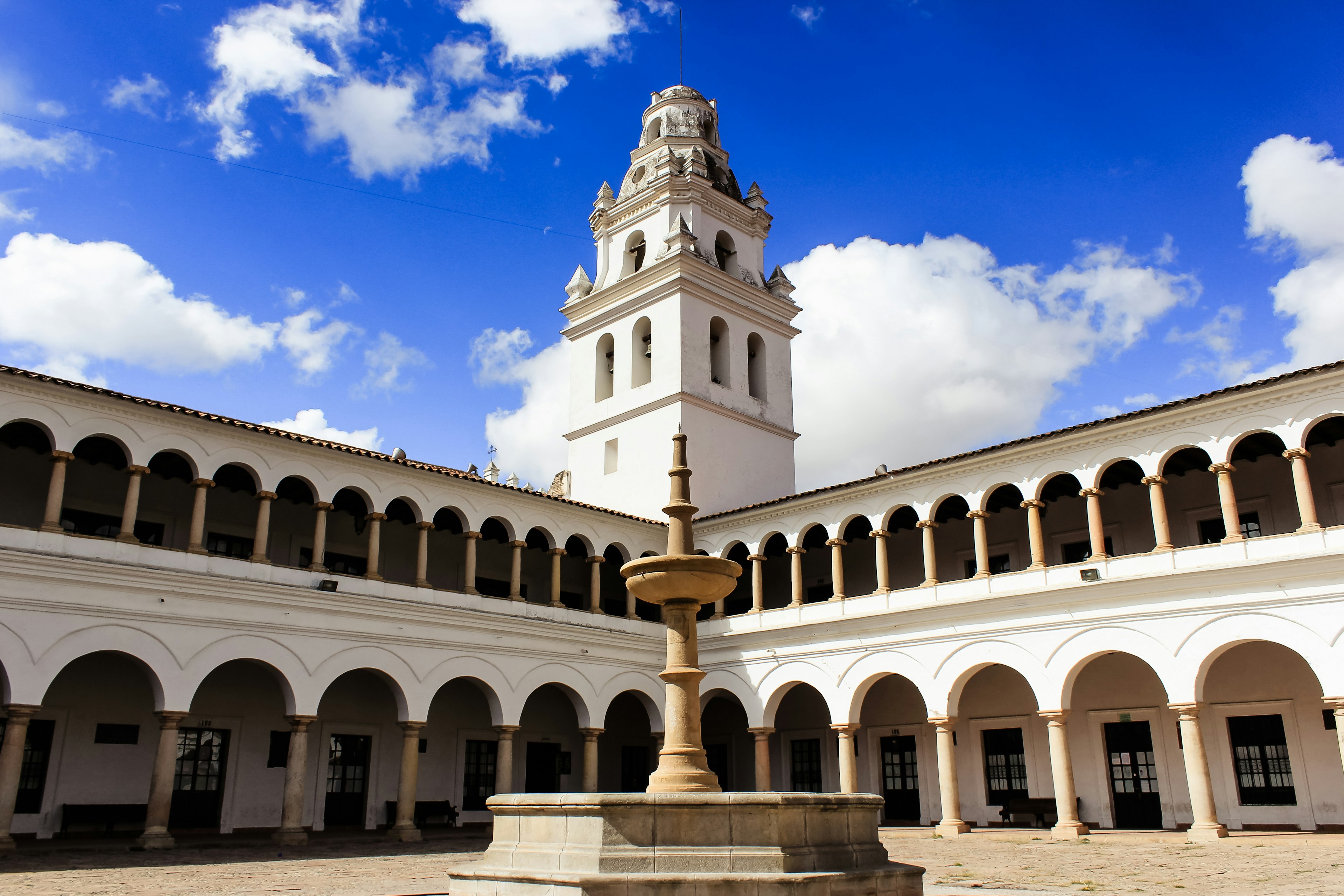 A fountain in a courtyard of a white building with symmetrical arched walkways and a tall bell tower