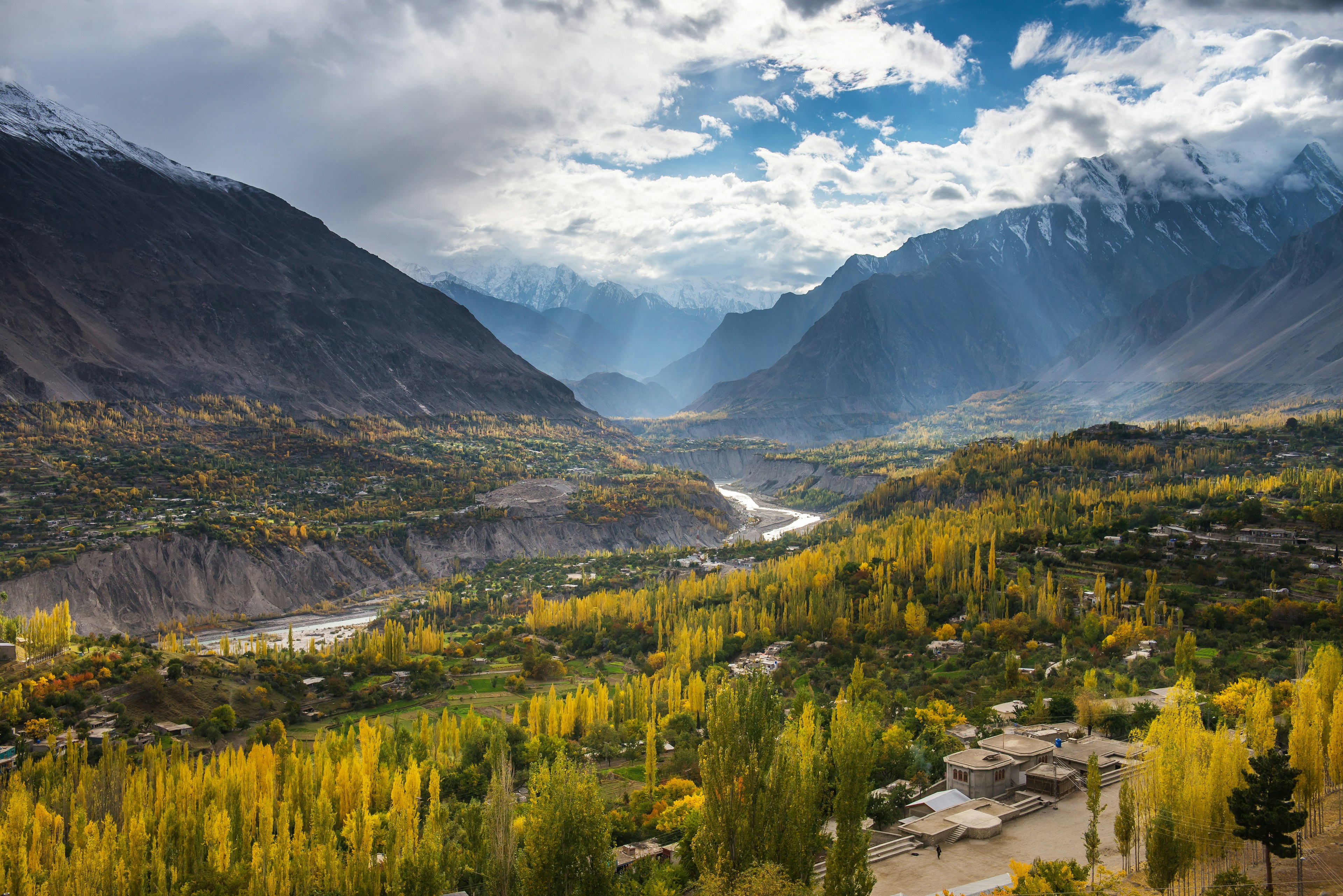 A river carves its way through a mountain valley lined with trees turning yellow in the autumn sun