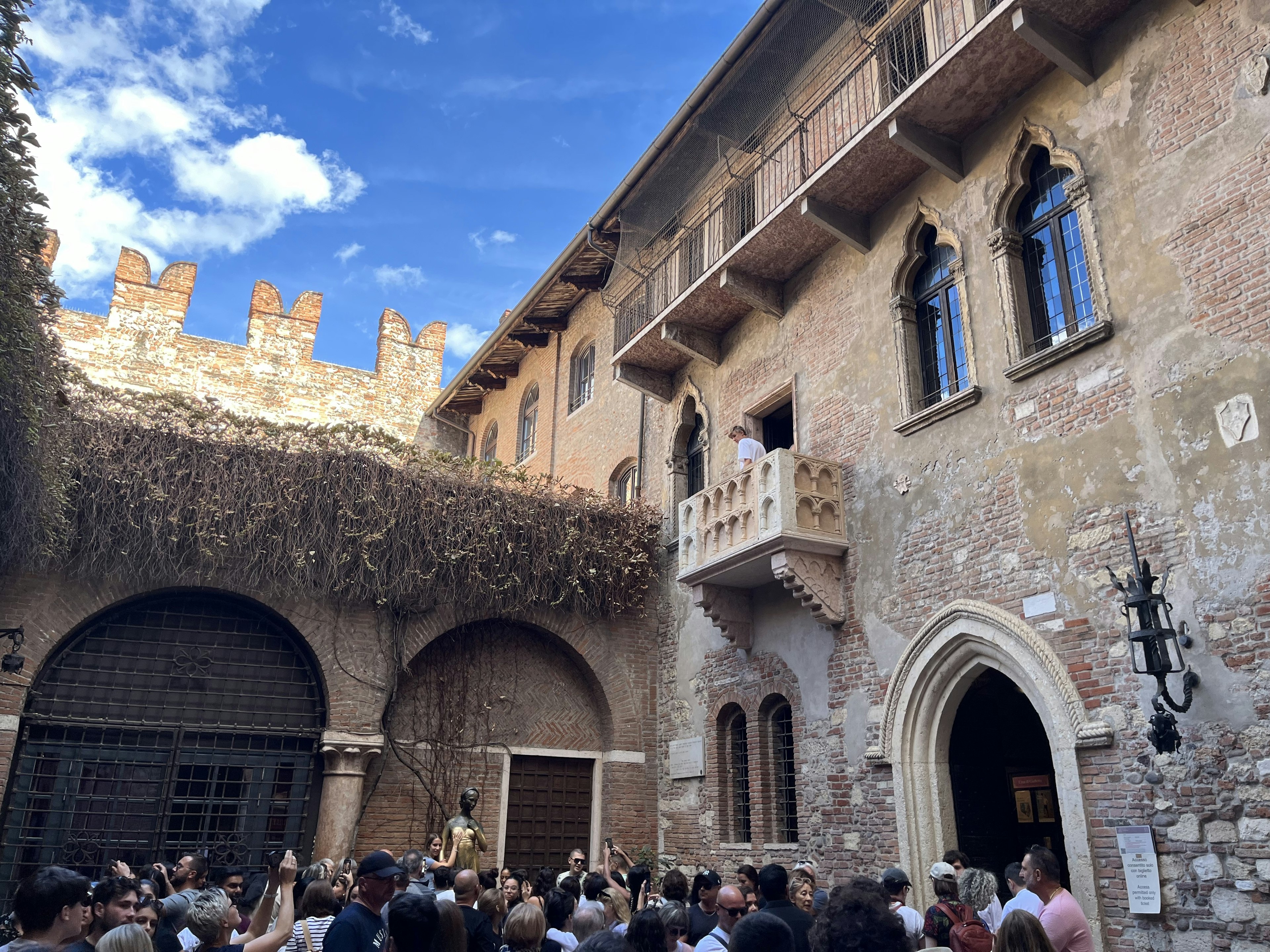 A person peers over a stone balcony to a crowd of people in a courtyard below