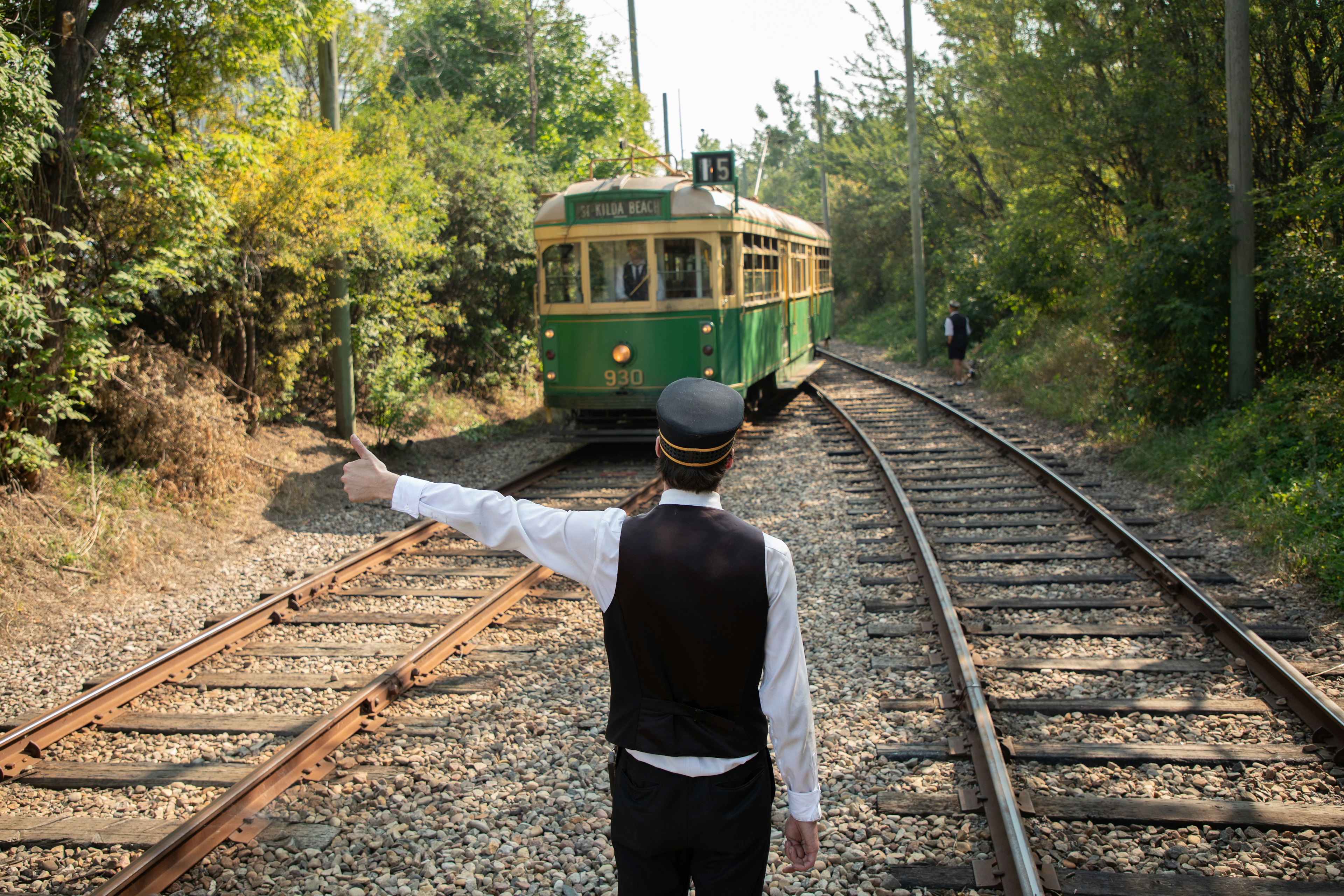 A conductor wearing a vintage uniform signals to a street car on the tracks, in a wooded area