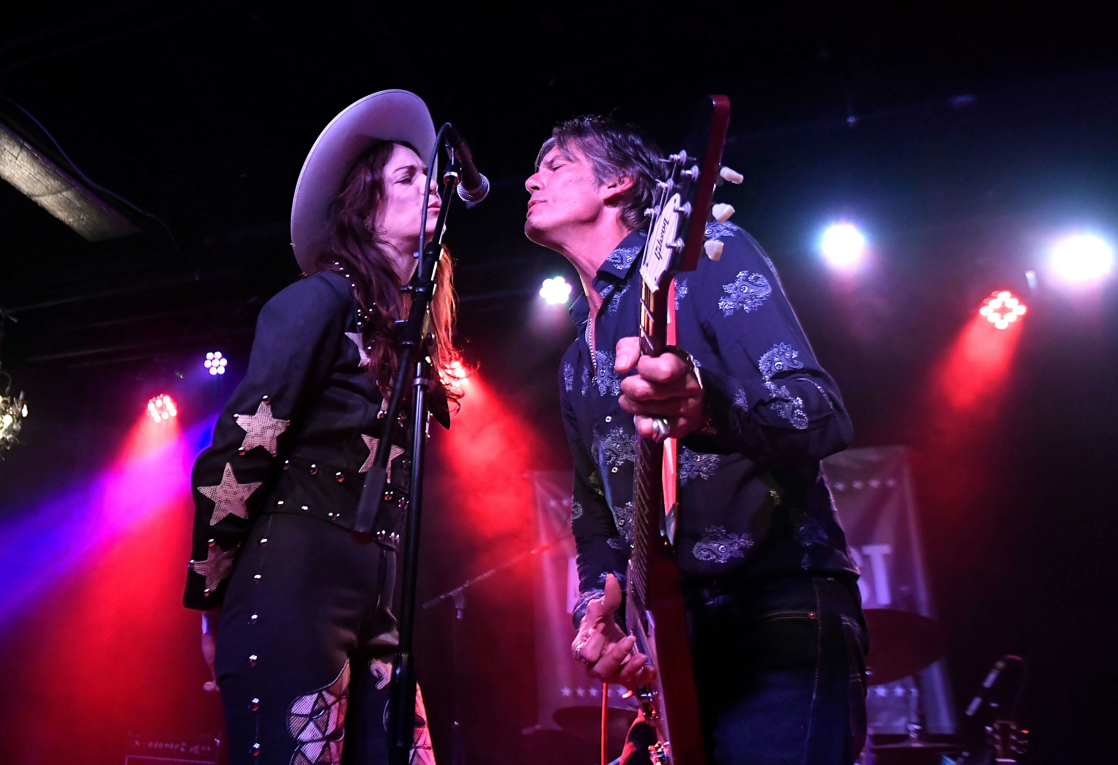 Two performers, one wearing a cowboy had and a shirt with star appliqués, the other playing a guitar, sing into a microphone on a concert stage lit by red stage lights