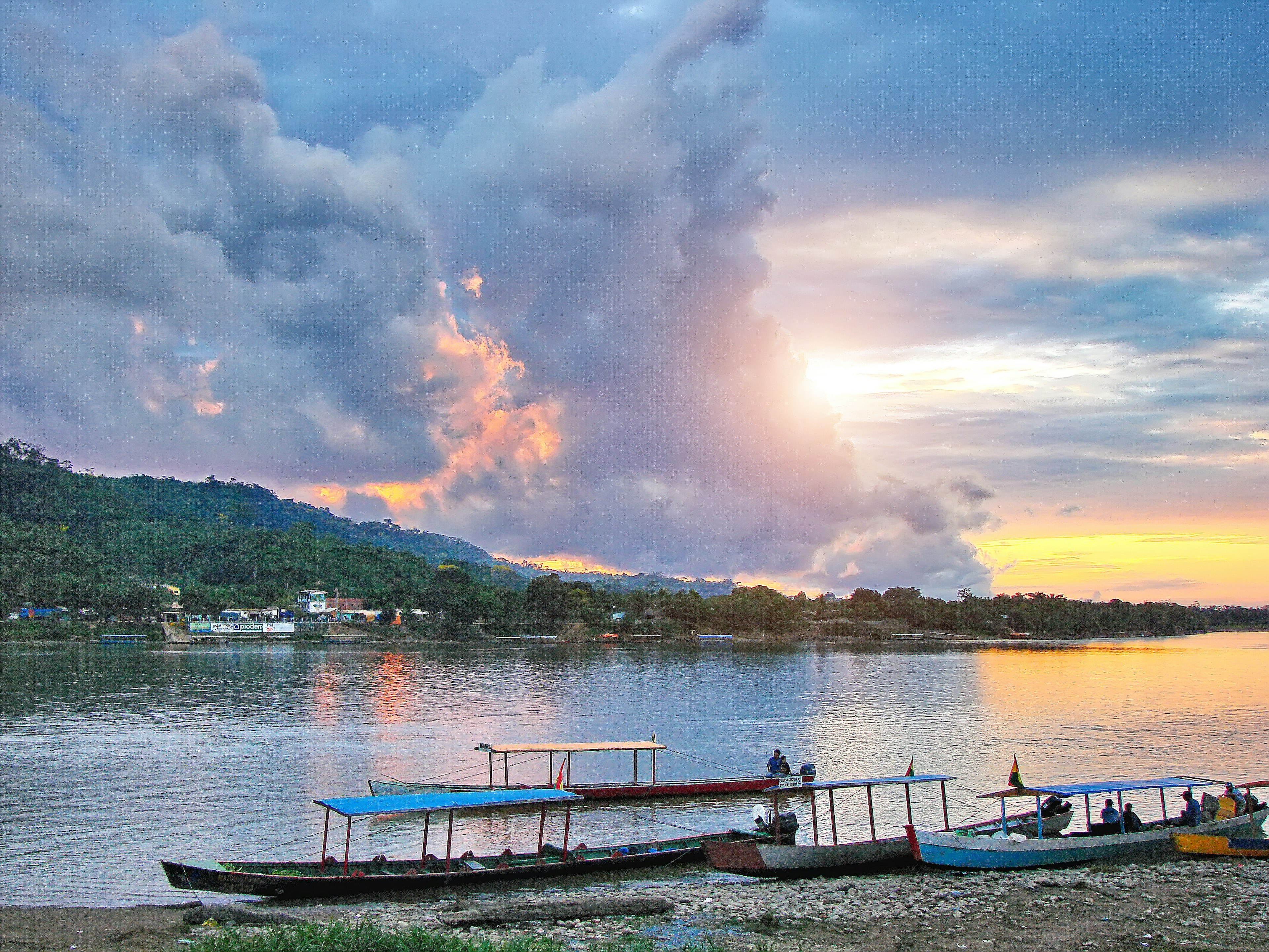 Long covered boats with benches docked at the side of a wide river in a remote jungle area at sunset