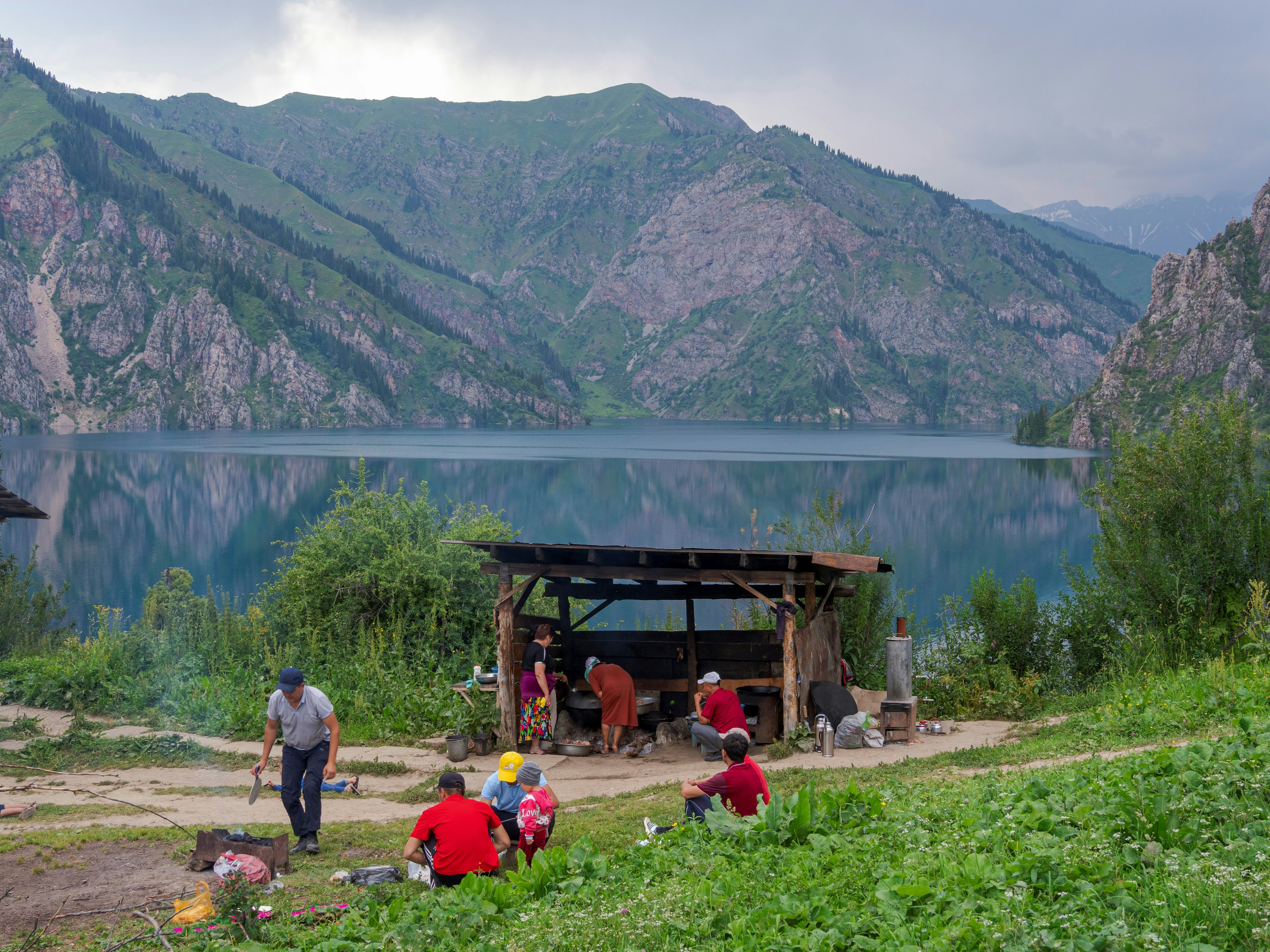 Local tourists prepare barbecue in a shelter by a lake with tree-covered slopes rising on all sides, Lake Sary-Chelek, Kyrgyzstan