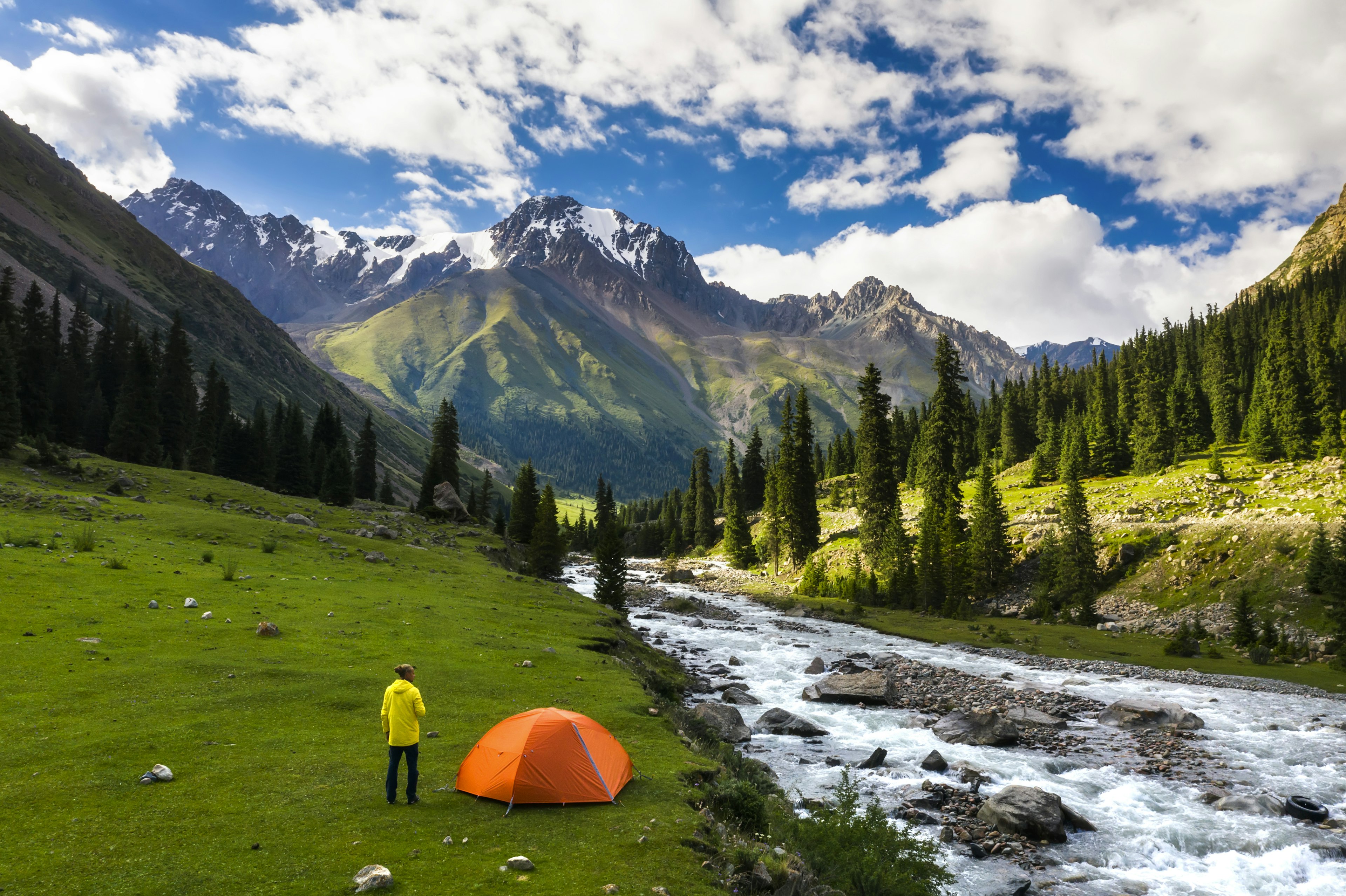 A man stands next to his orange tent in a green mountain valley next to a roaring river. Snow-capped peaks and pine trees are visible in the distance.