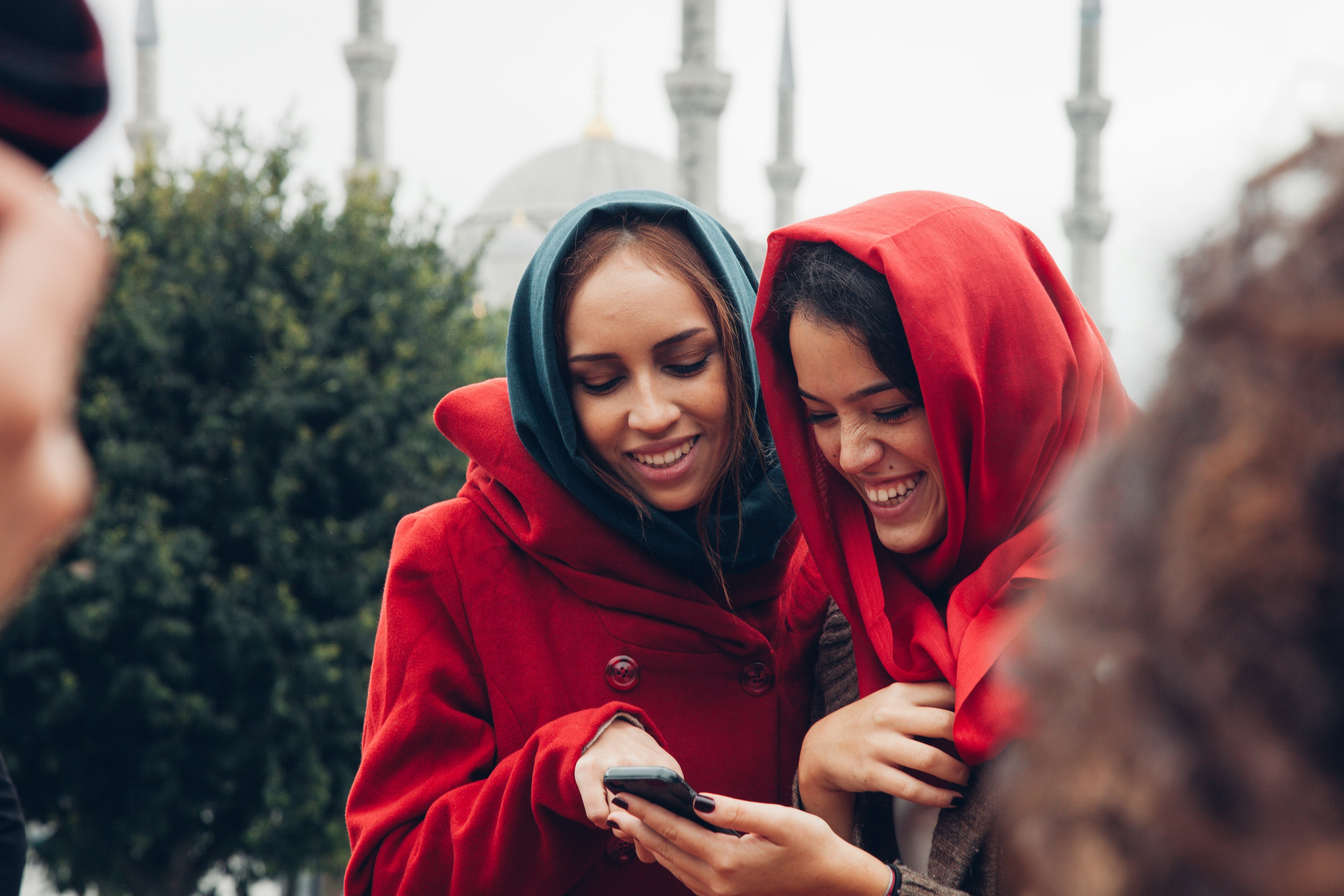 Two young women in head coverings laugh as they look at a phone near the Blue Mosque in Istanbul, Türkiye