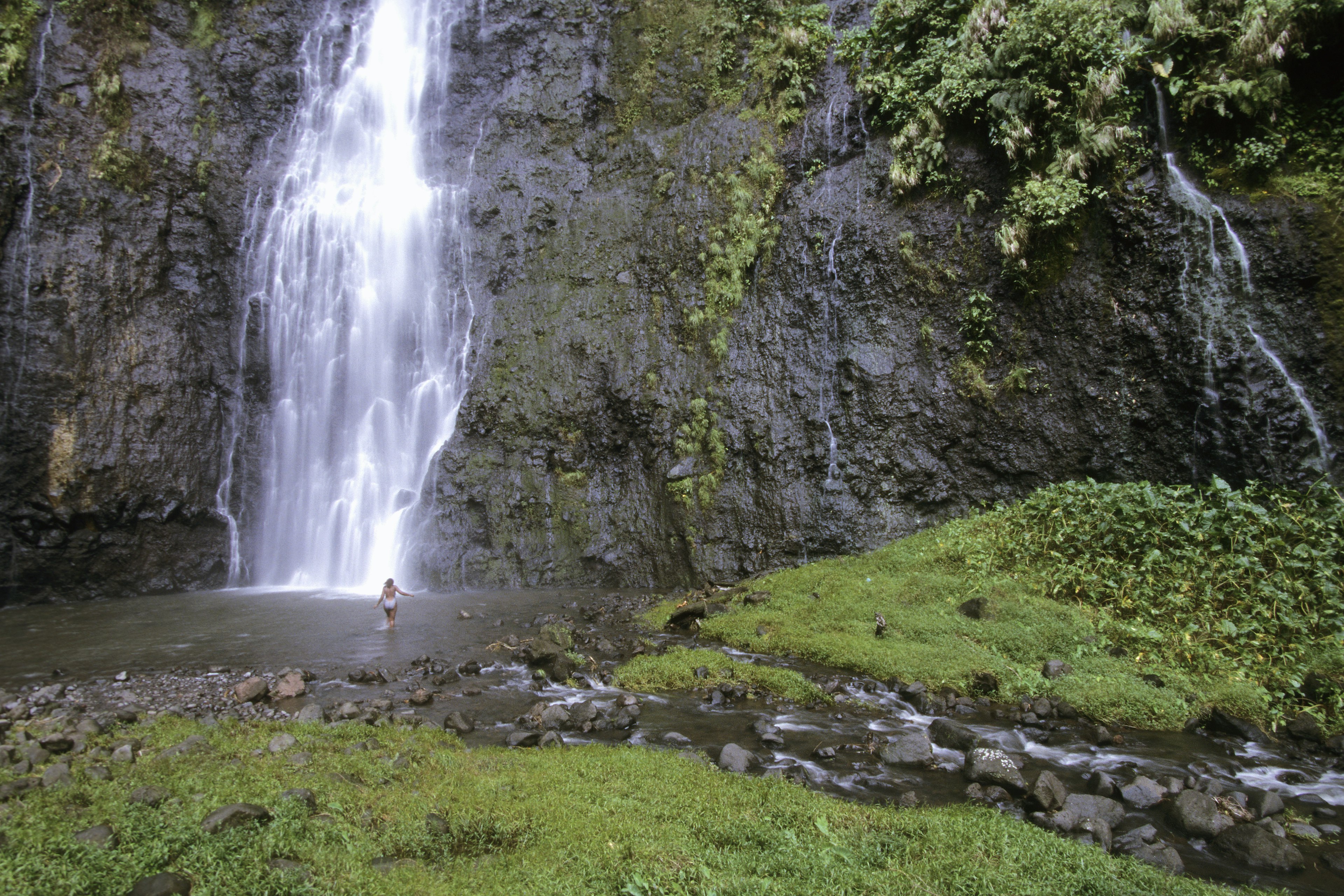 A woman wades into a cascading waterfall in the jungle.