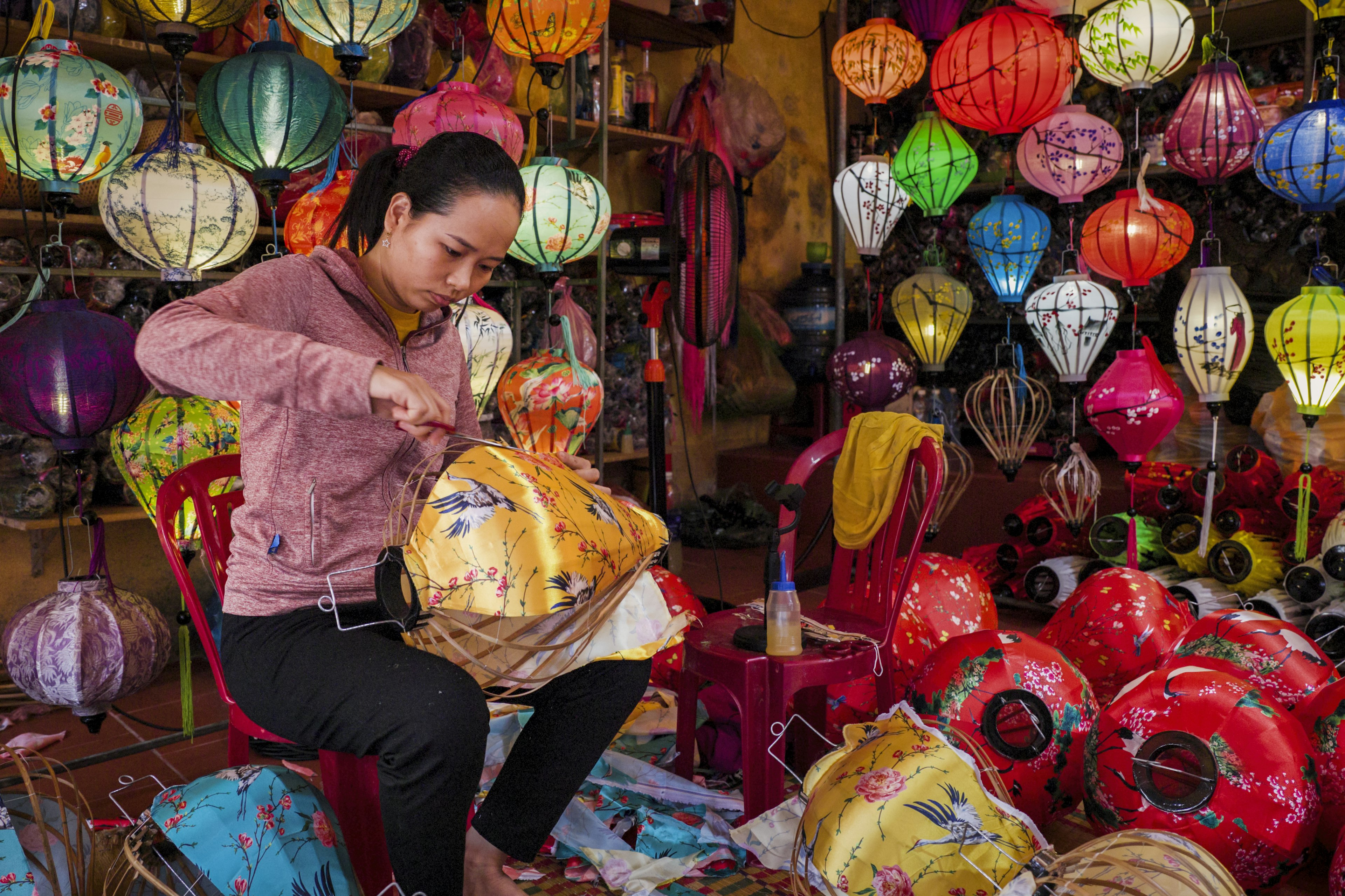 A woman crafts an orange lantern in a workshop filled with other colorful lanterns in various stages of manufacture