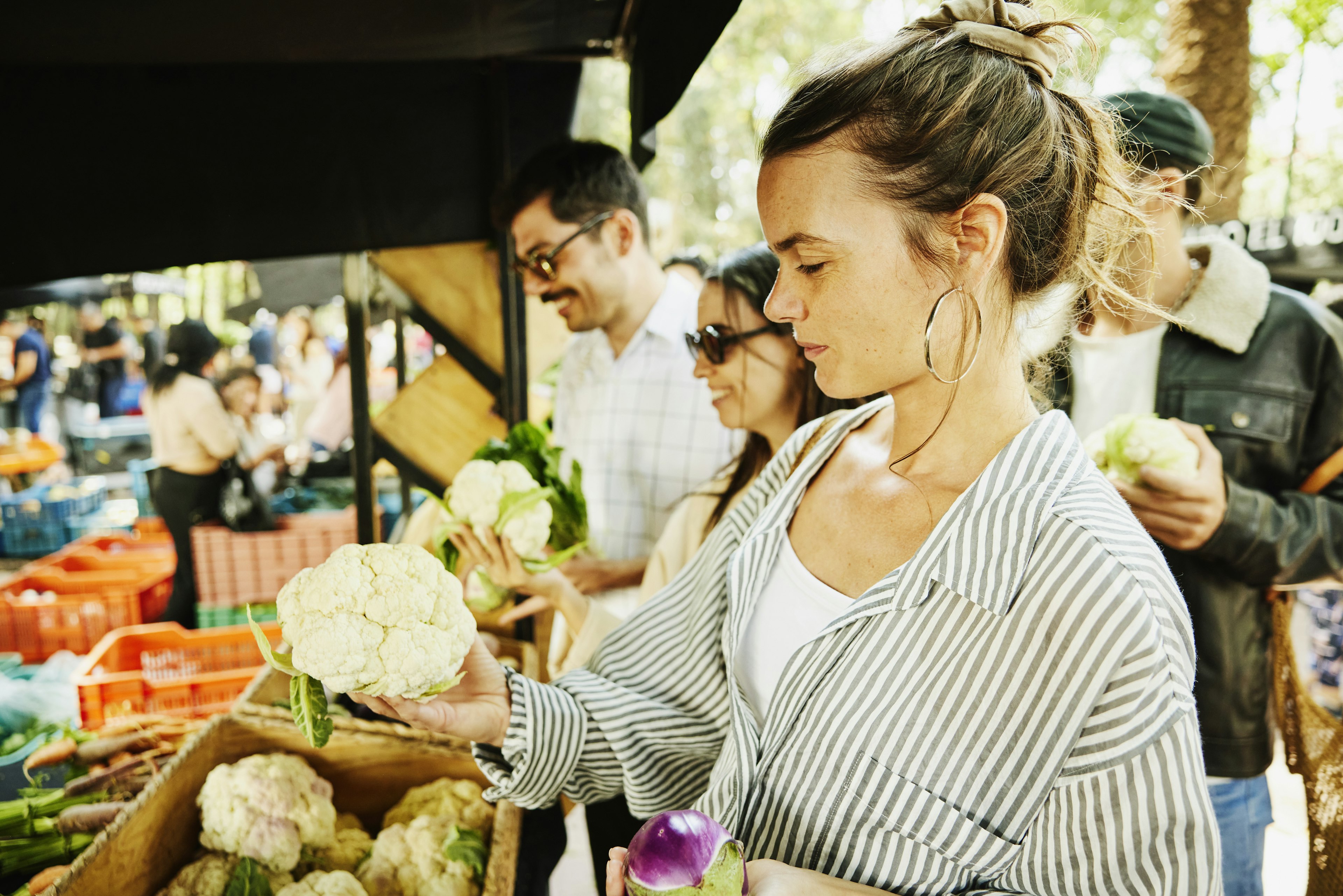 Medium shot of woman in striped shirt selecting cauliflower while shopping with friends at organic farmers market