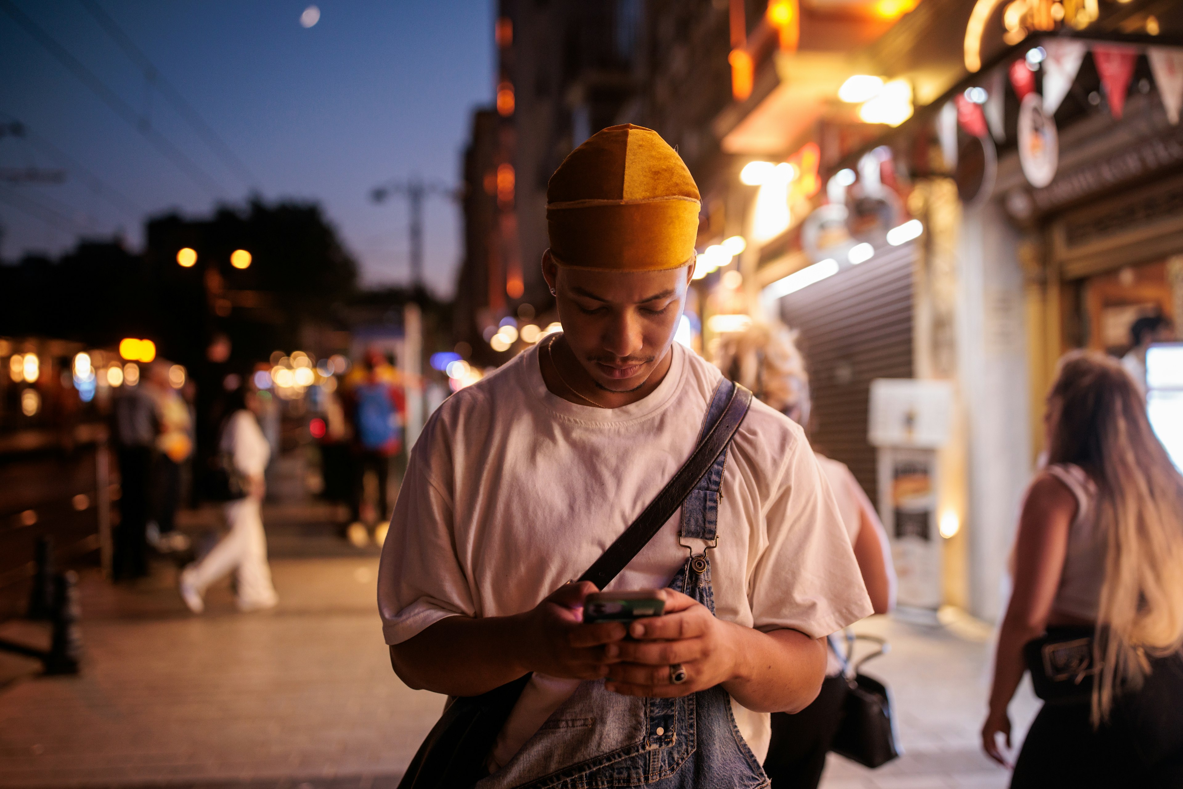 A male tourist in a white t-shirt and overalls checks his phone at night on a busy street in Istanbul, Türkiye