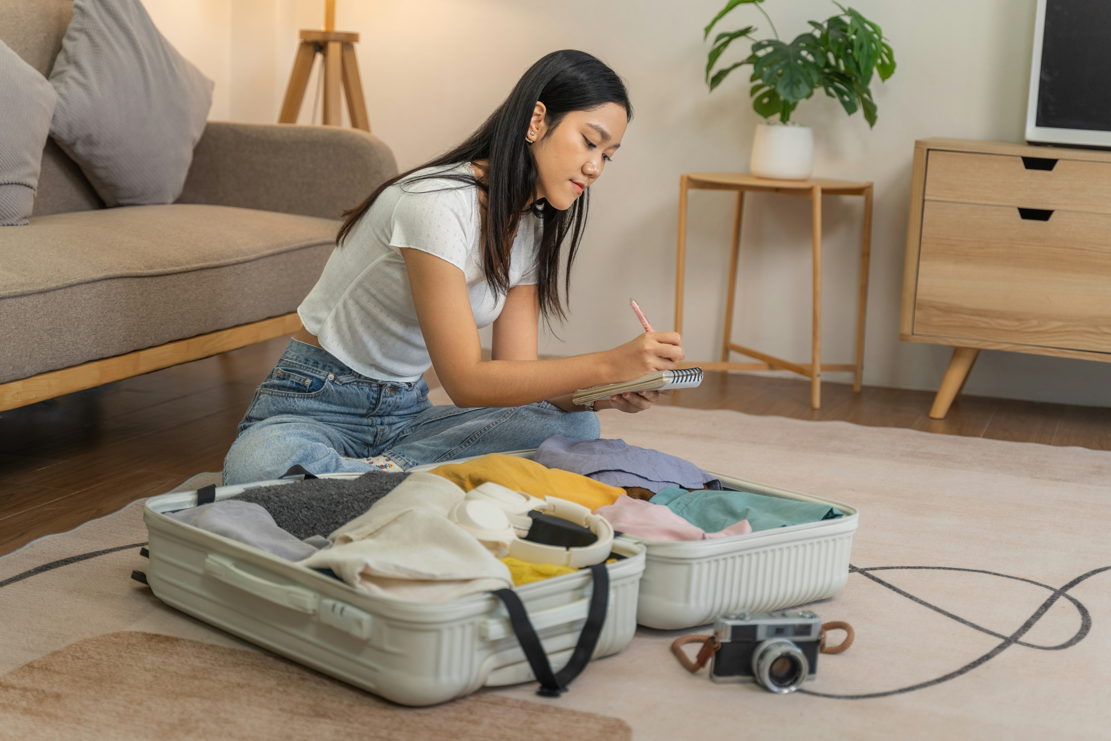 A female is seated on the floor of her living room checking her list on her notepad while packing her suitcase.