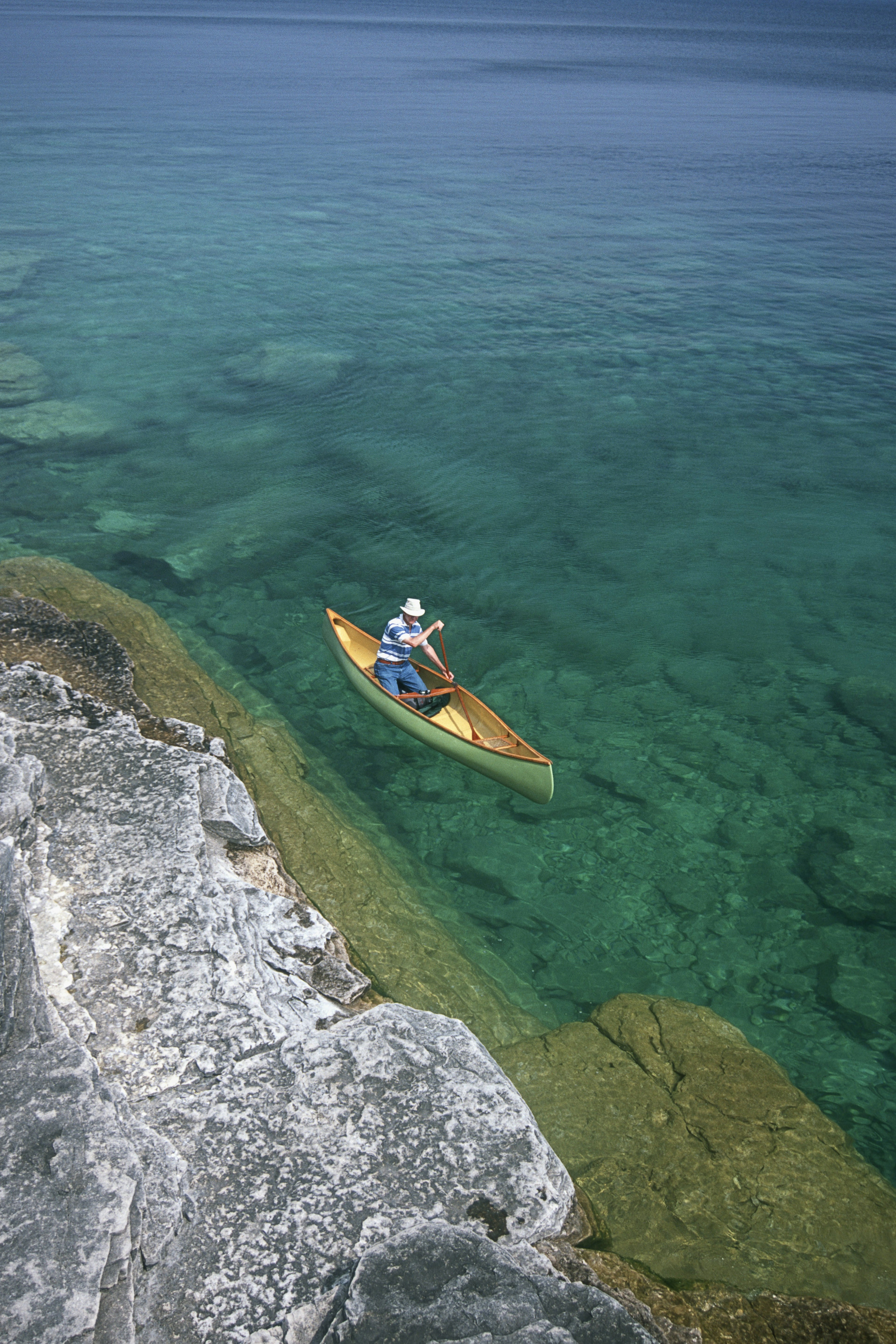 A single paddler canoes along the rocky shoreline; taken from above, the waters around the canoe are emerald in colour and the bottom is clearly visible through the transparent water.