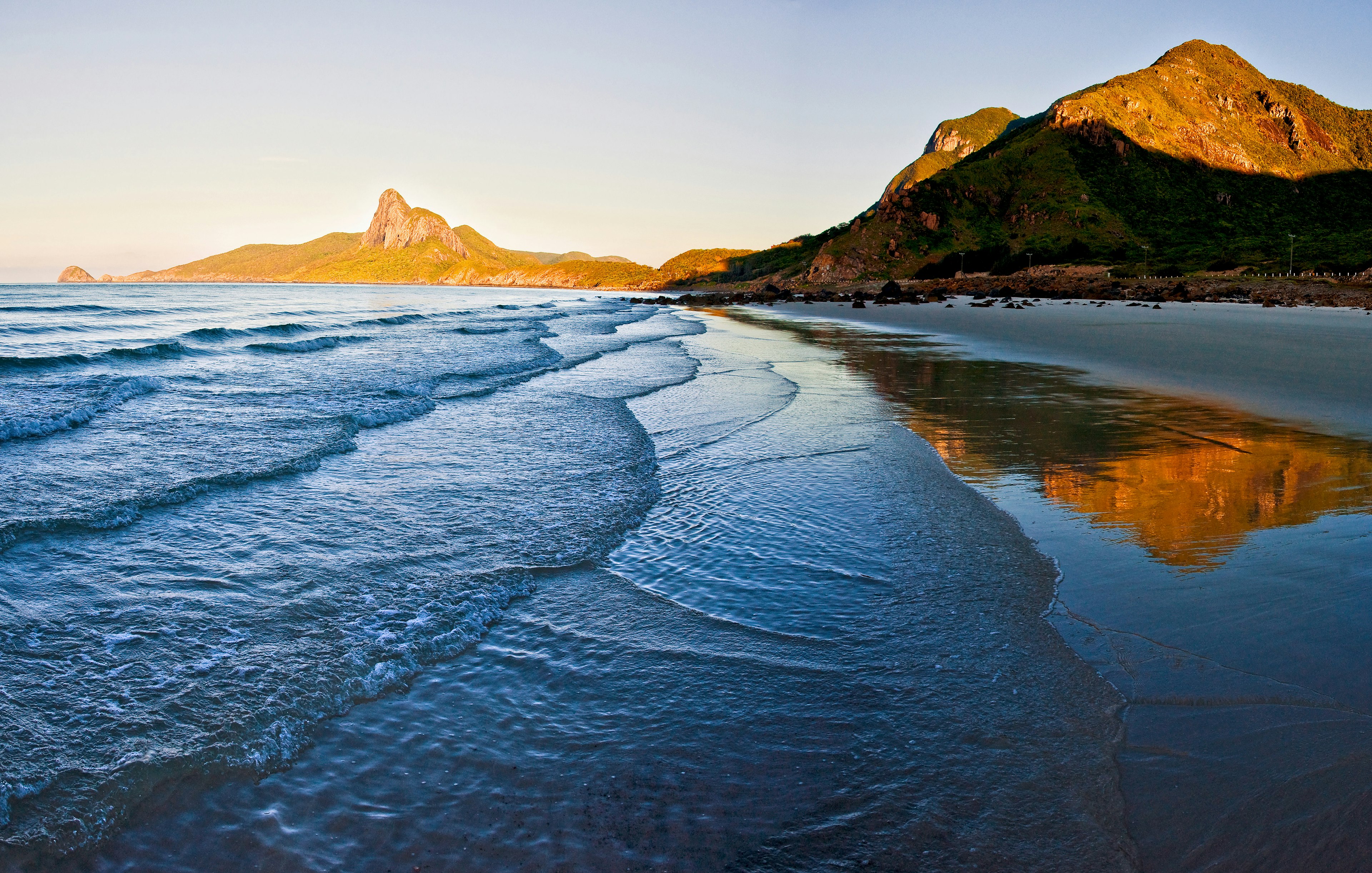 Gentle waves lap the shore at sunrise on a beach on Con Dao Island. Rock-faced mountains bathed in morning light can be seen in the distance.