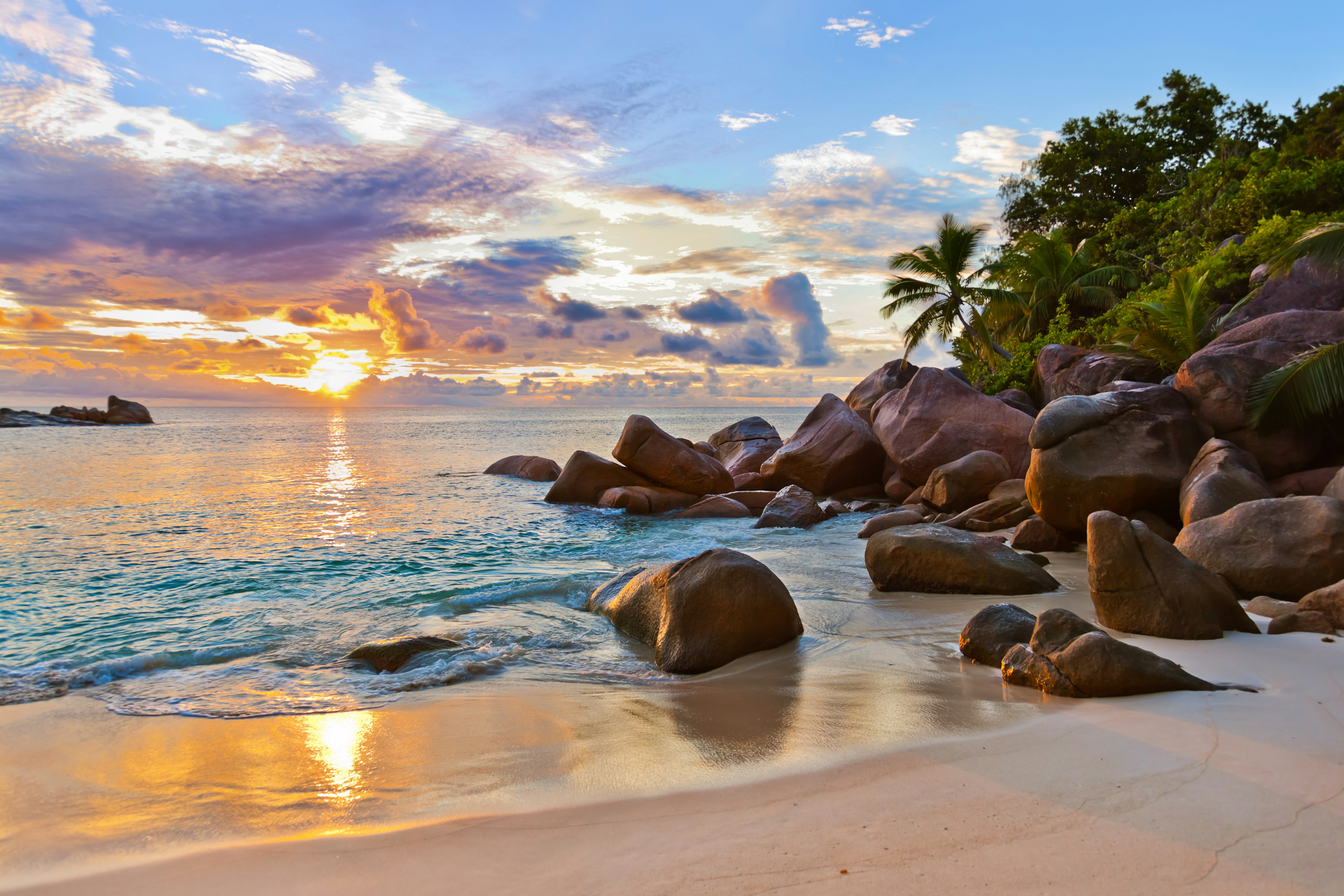 A beautiful cove in the Seychelles; the sky is a dramatic canvas of clouds and the setting sun, while the sea is greeny-blue and surf laps at a golden shore; at the end of the beach, large grey rocks rise to meet palm trees and other greenery.