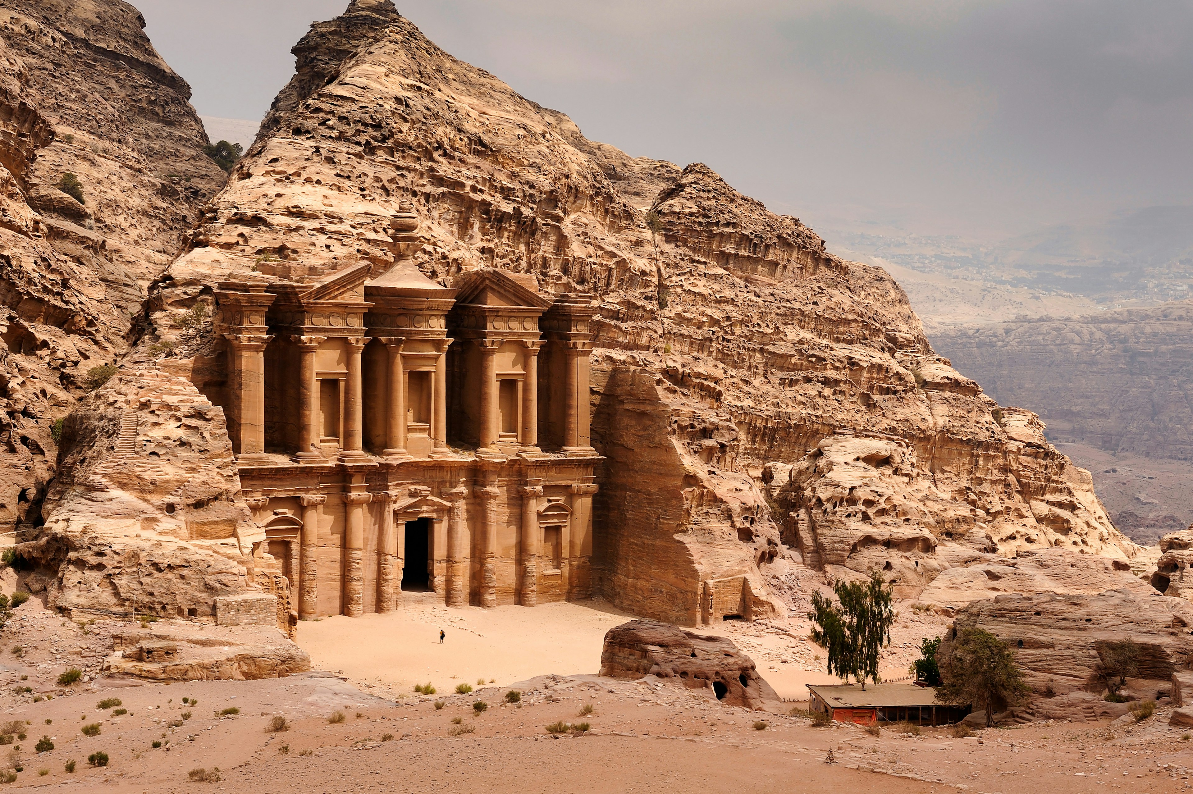 A person stands near a doorway to a massive building built and carved into stone in a hilly desert area