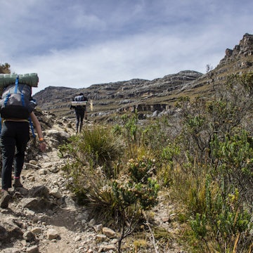 Two hikers, both wearing huge backpacks complete with sleeping mats, climb up a rocky trail within the mountains; the scene is quite barren, with no trees, just low shrubs and rock outcrops.
