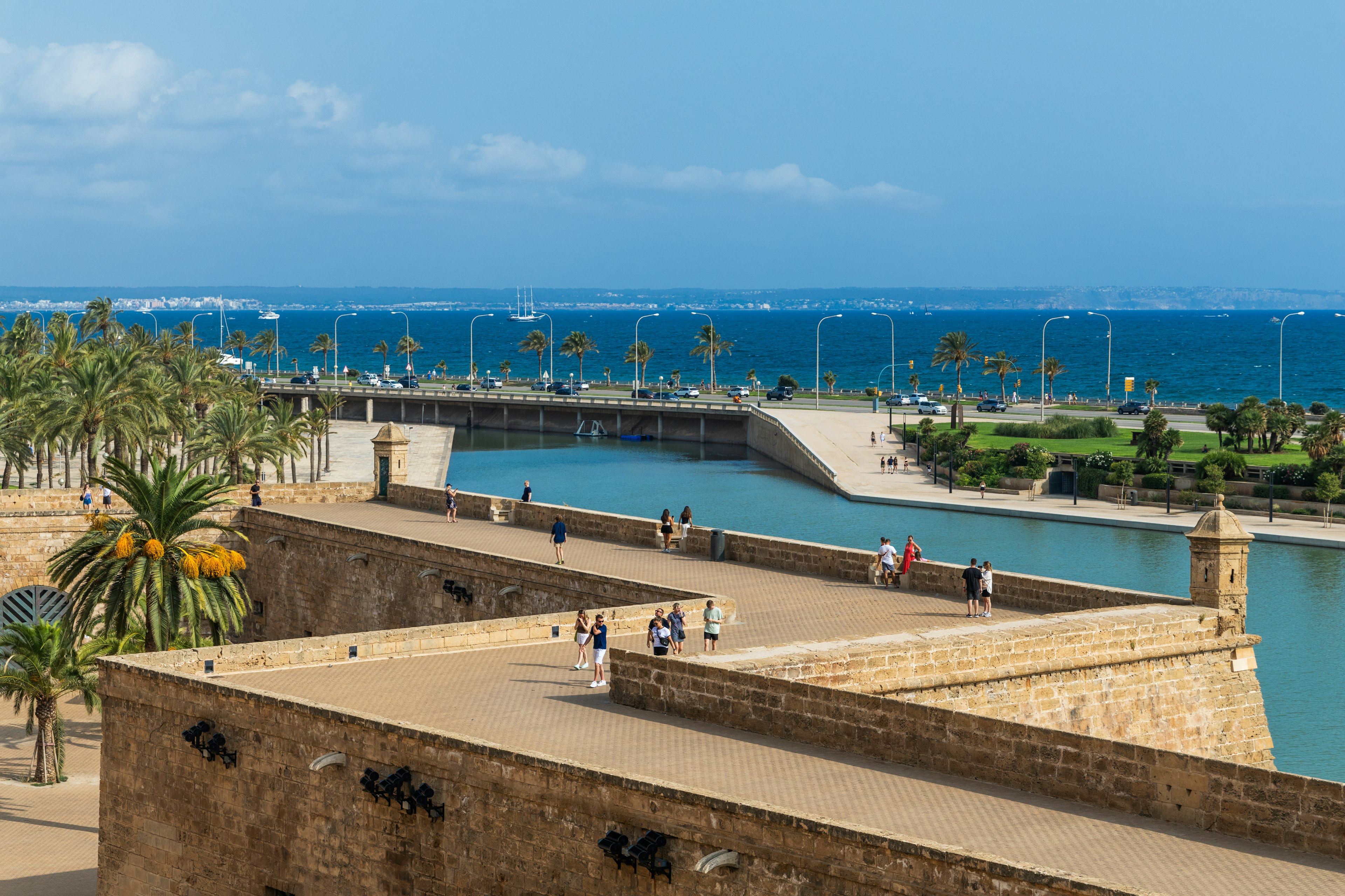 Tourists walking along the stone walls of the Almudaina Palace overlooking the waterfront in Palma.