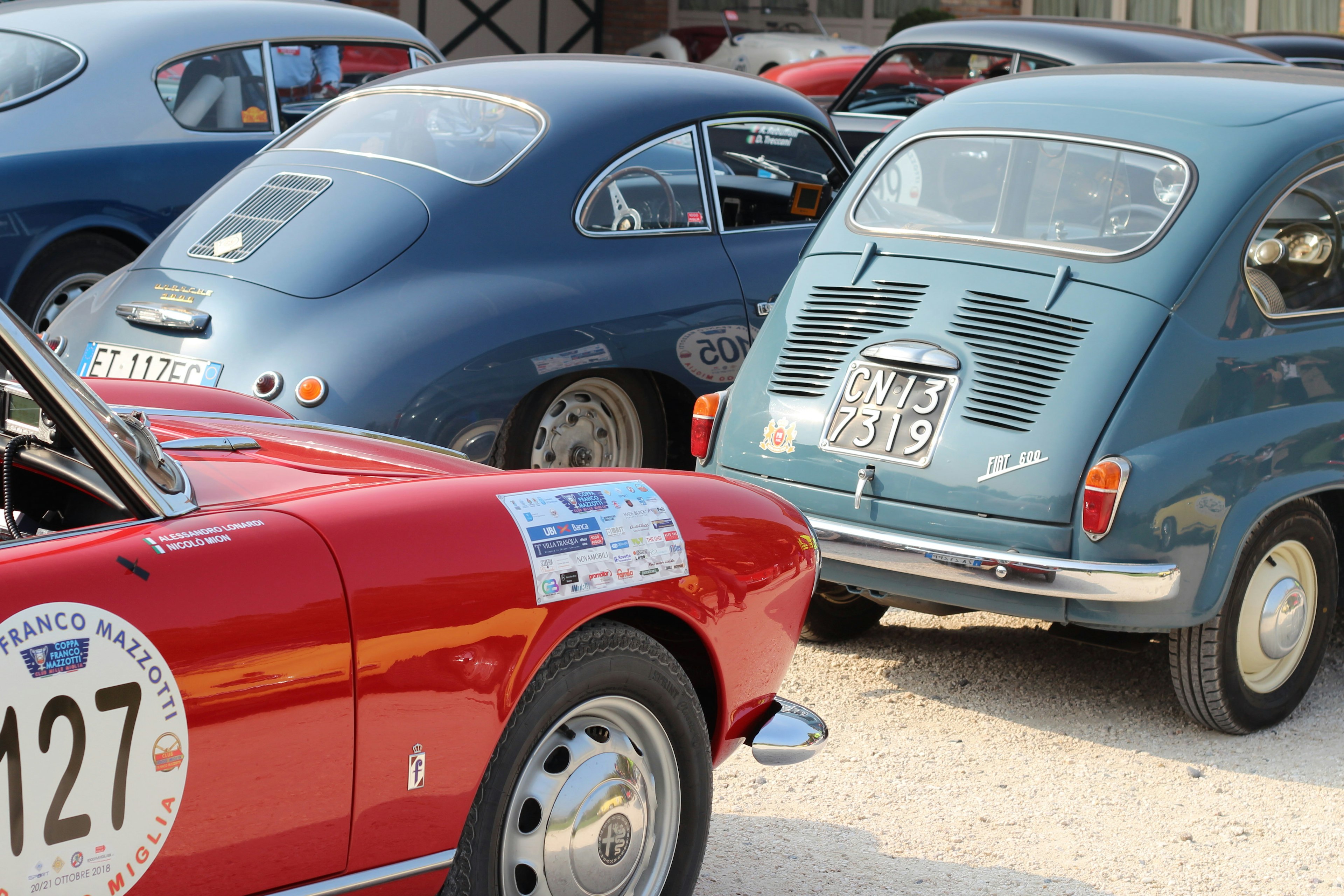 Red British convertible car, a 1955 AC Ace, with blue Fiat 600 and Porsche 4800 in background at the Coppa Franco Mazzotti classic car rally.