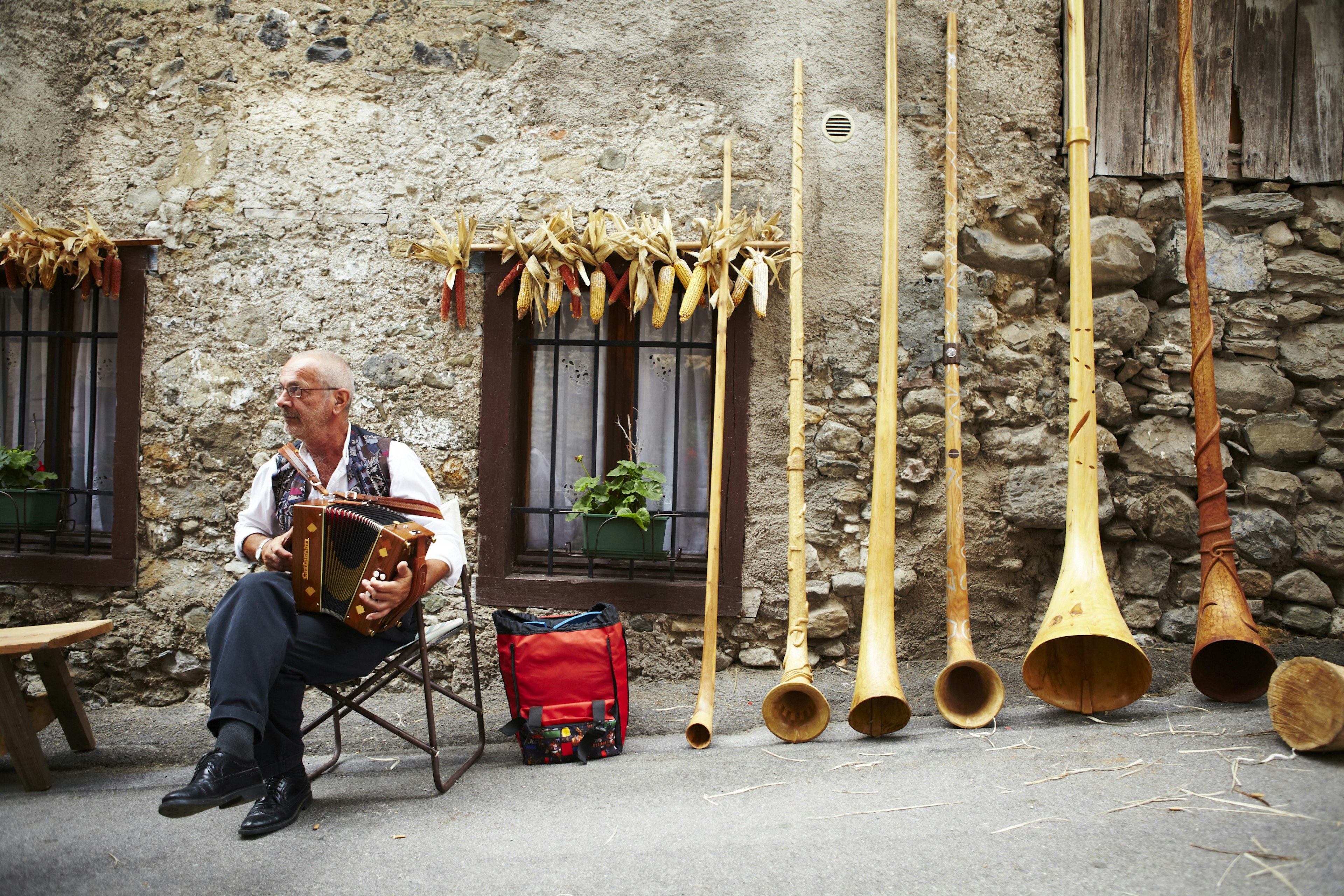 A man in a traditional embroidered vest sits in a chair and plays an accordion in a village street. Several large alpenhorns lean against a stone wall to the musician’s left.