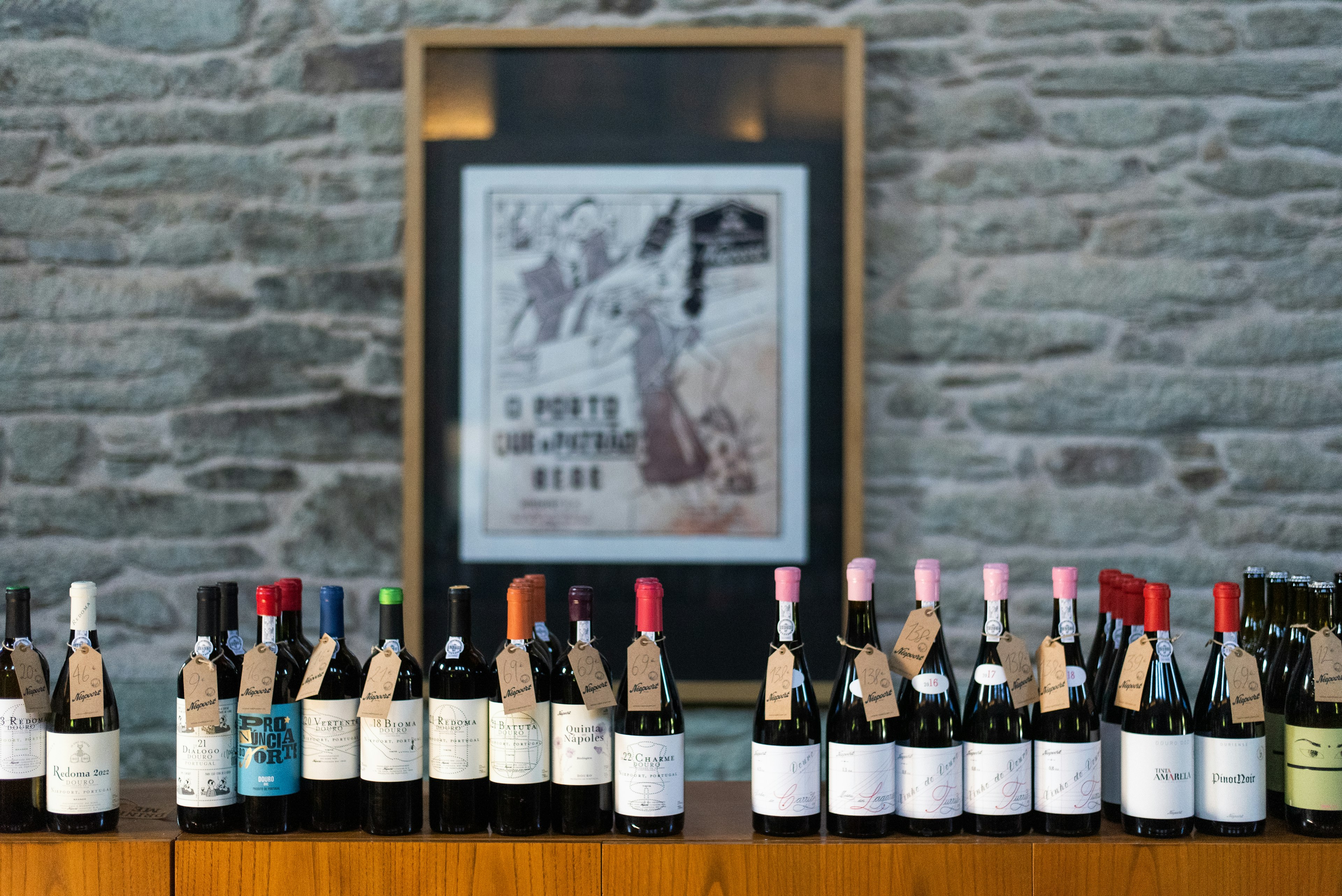 A series of wine bottles with paper labels is lined up on a counter, in front of a stone wall with a framed poster