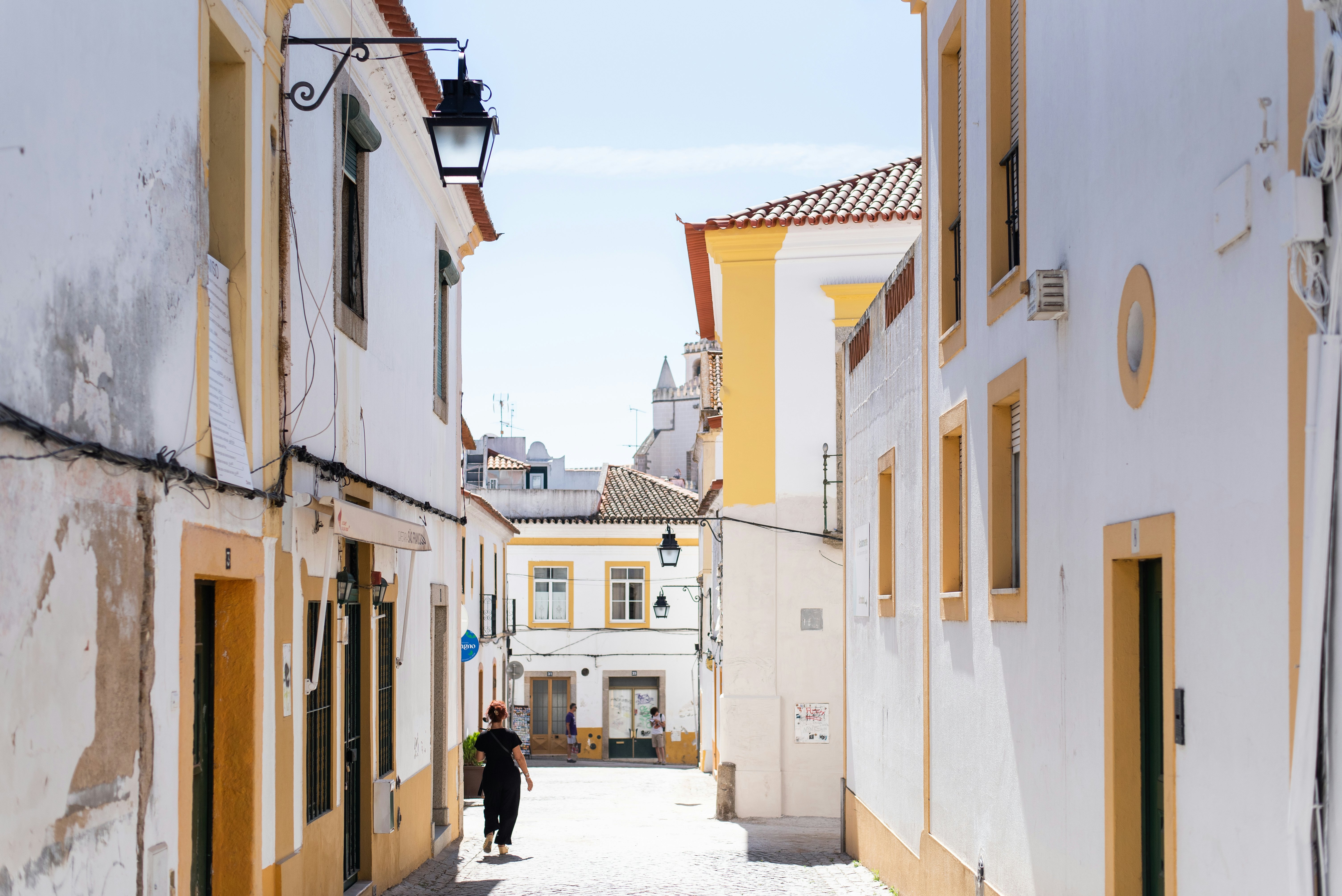 A woman walks down a street with white buildings with yellow-trimmed windows and doors.