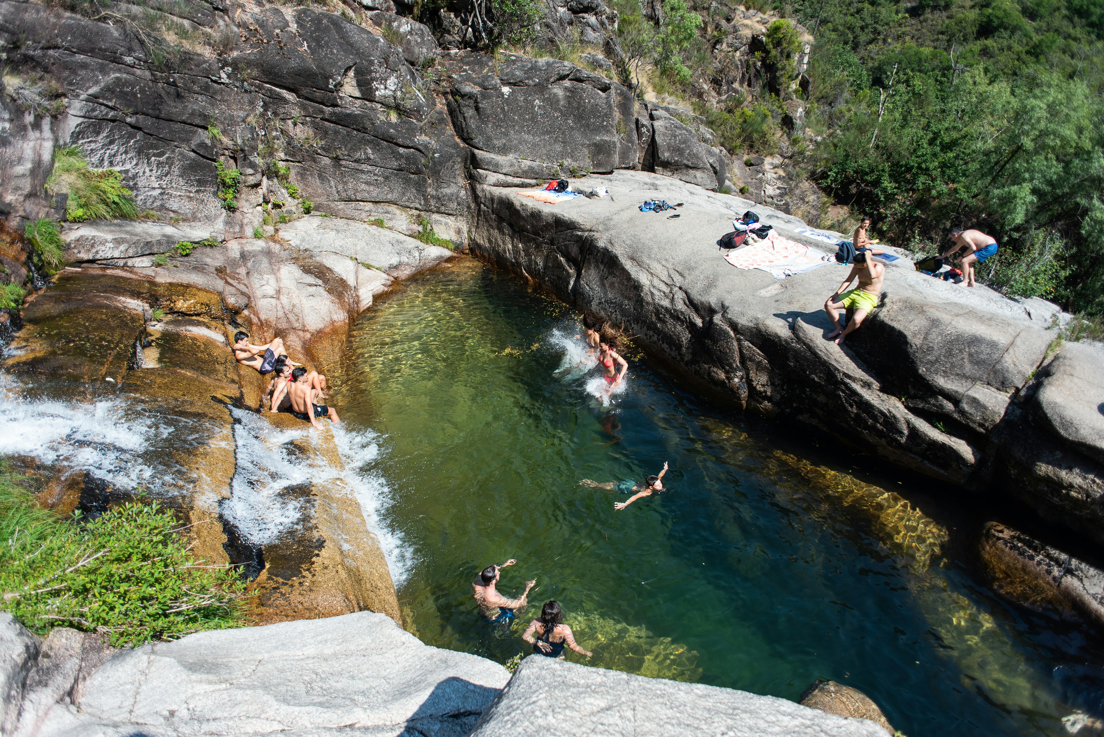 A group of people in bathing suits sit on rocks and swim in a pool fed by a waterfall on a sunny day