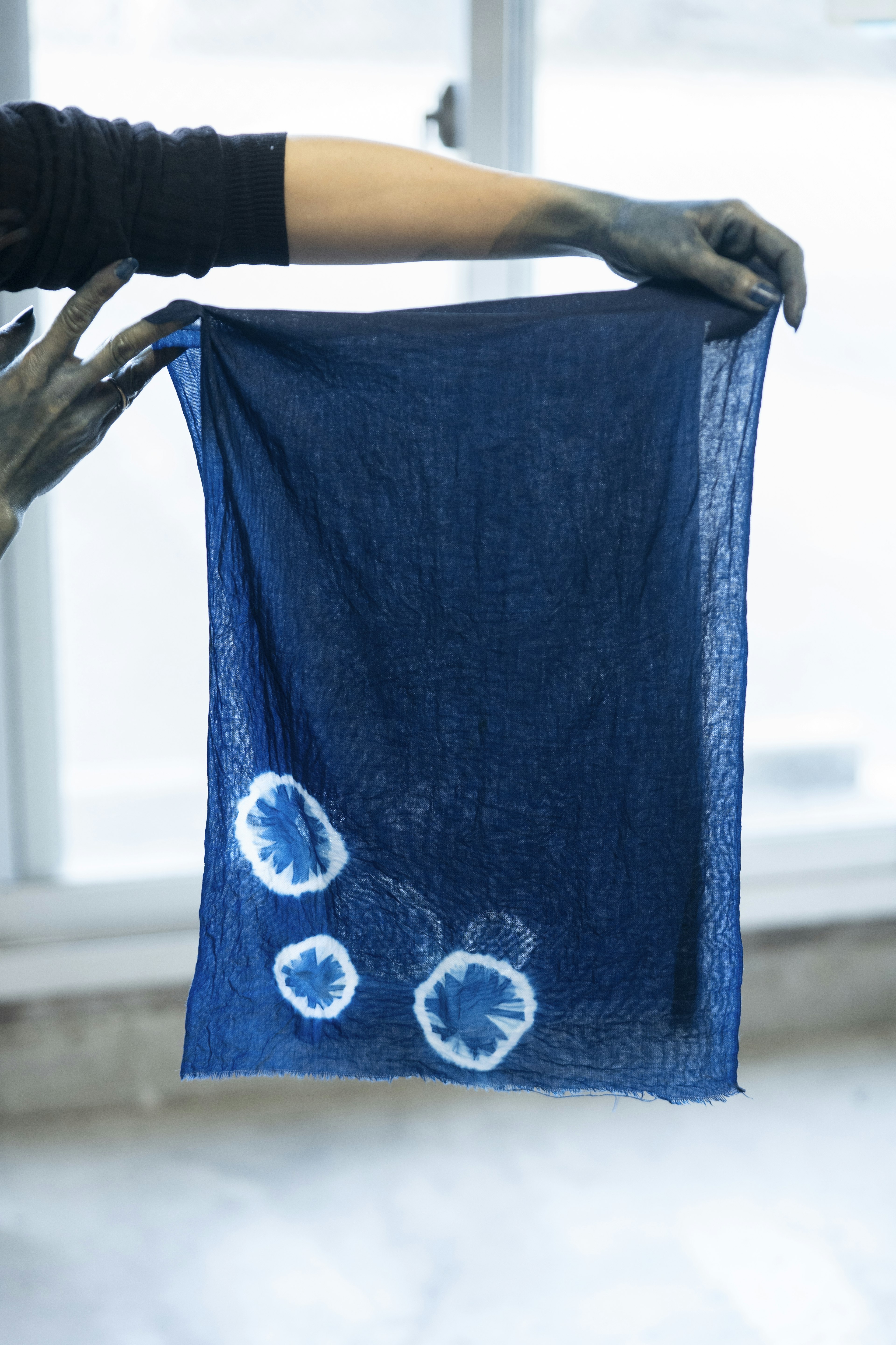 A person whose hands have been stained by dye holds up a piece of fabric that has been colored blue by hand