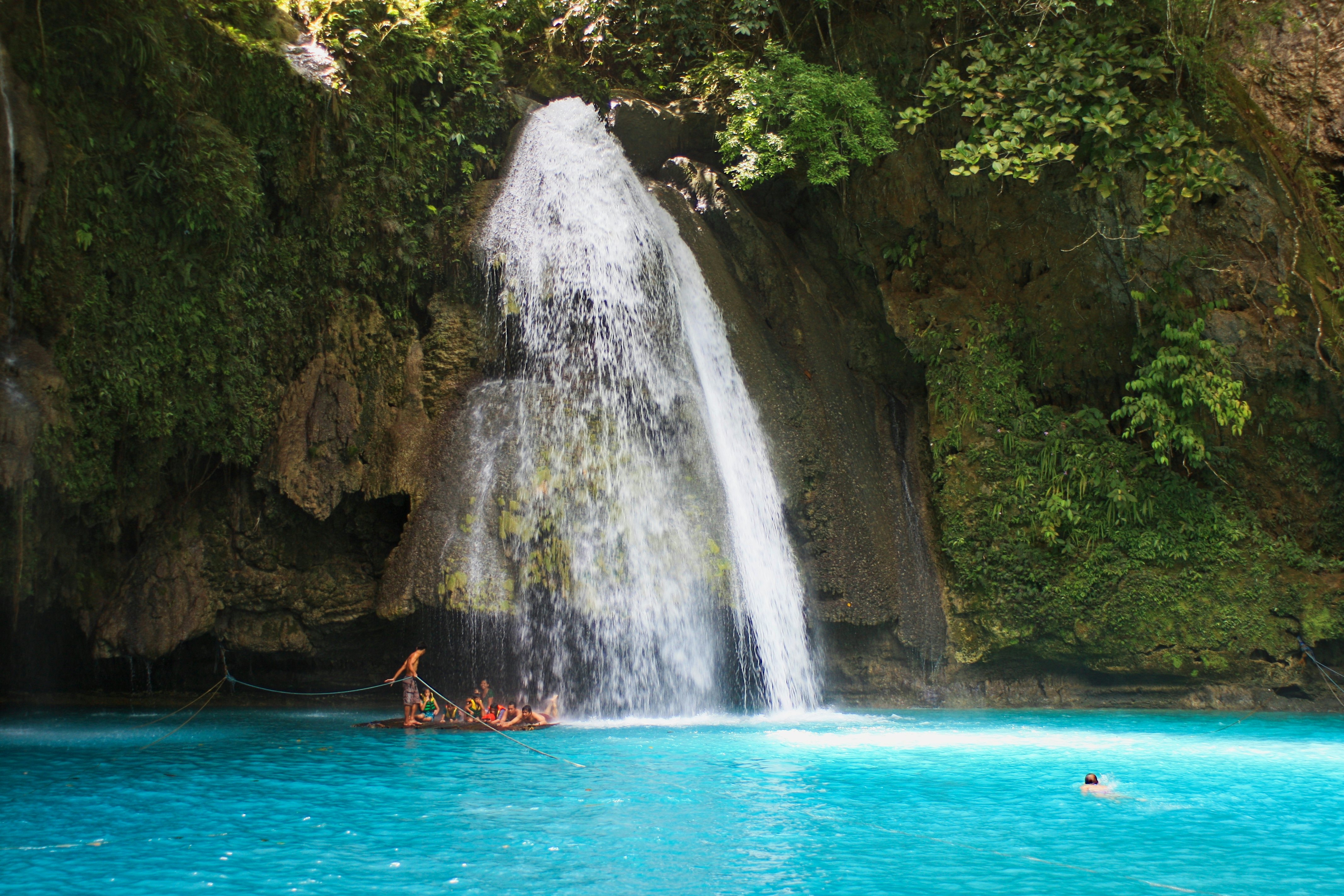 People swim in a blue lagoon at the base of a tall waterfall, which falls down a slope covered with greenery and rocks