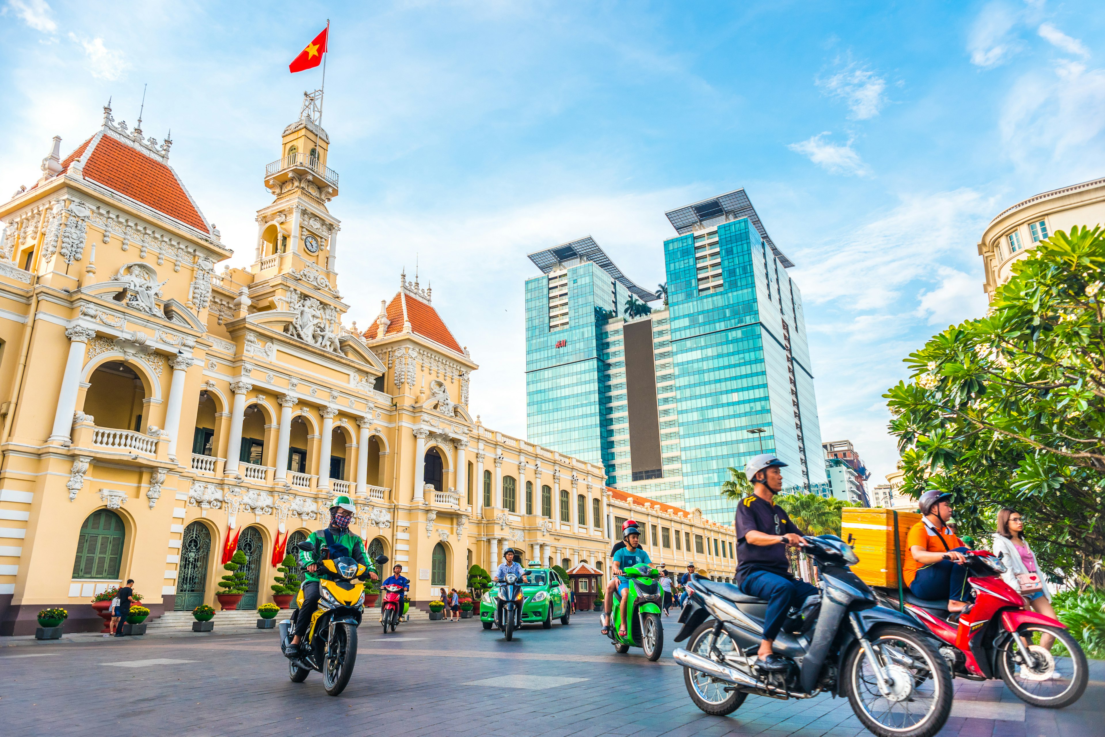 Motorcyclists pass by a large city hall building with a tower flying a red flag with a yellow star