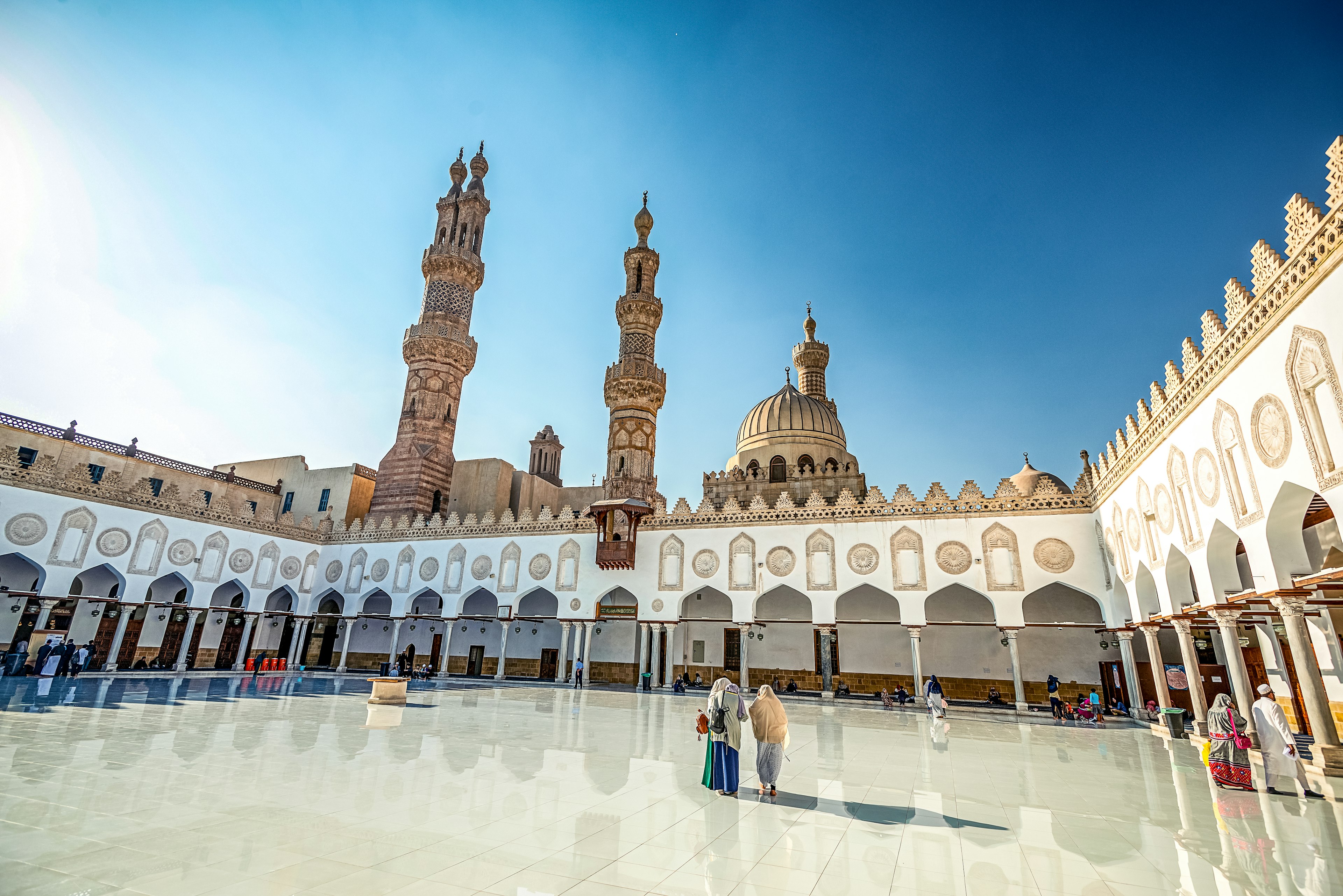 People walk across the central courtyard of a vast mosque complex with three tall minarets and a dome