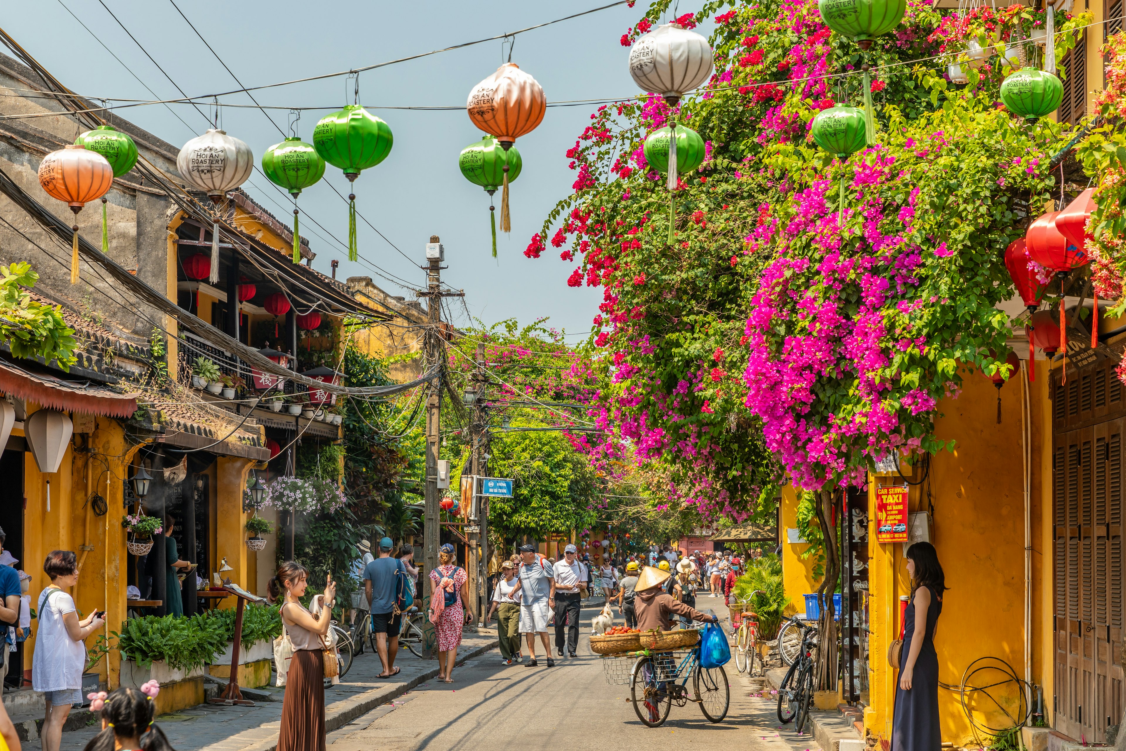 A busy street lined with plants as people passs through under lanterns that hang above