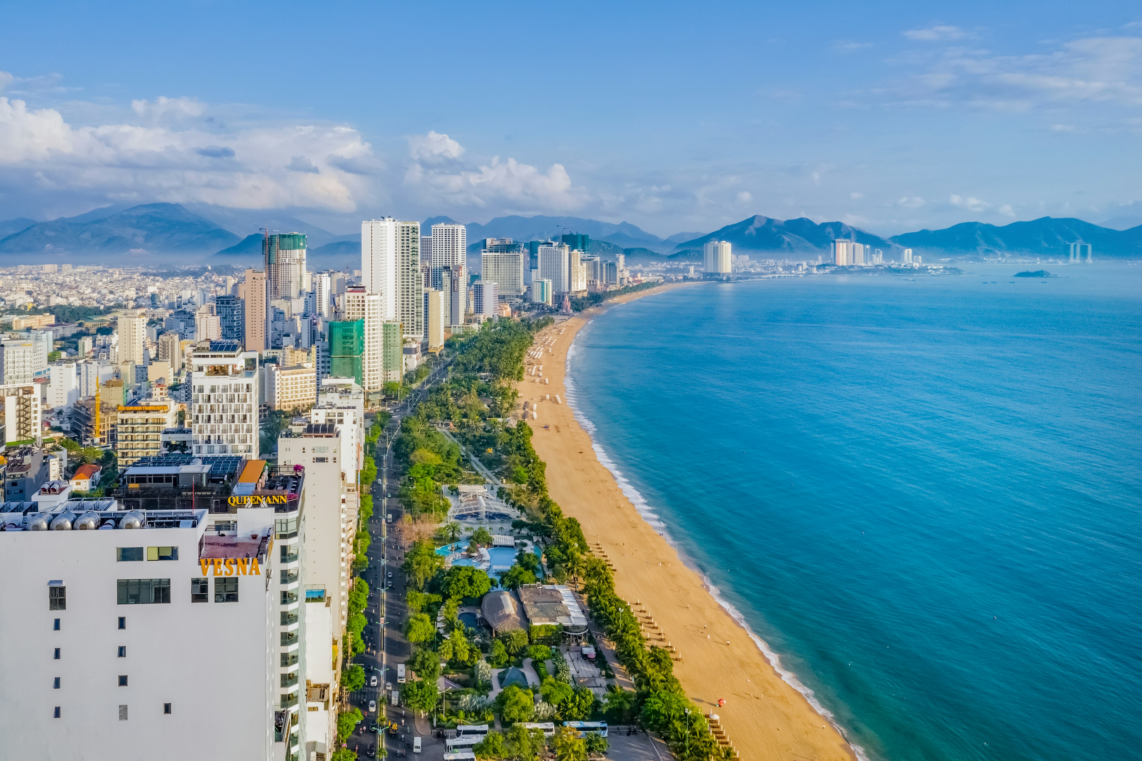 A wide aerial view of the city of Nha Trang, with a number of high-rise buildings on the left side of the shot and the golden strip of beach running alongside the city on the right-hand side, with the South China Sea in the right side of the frame.