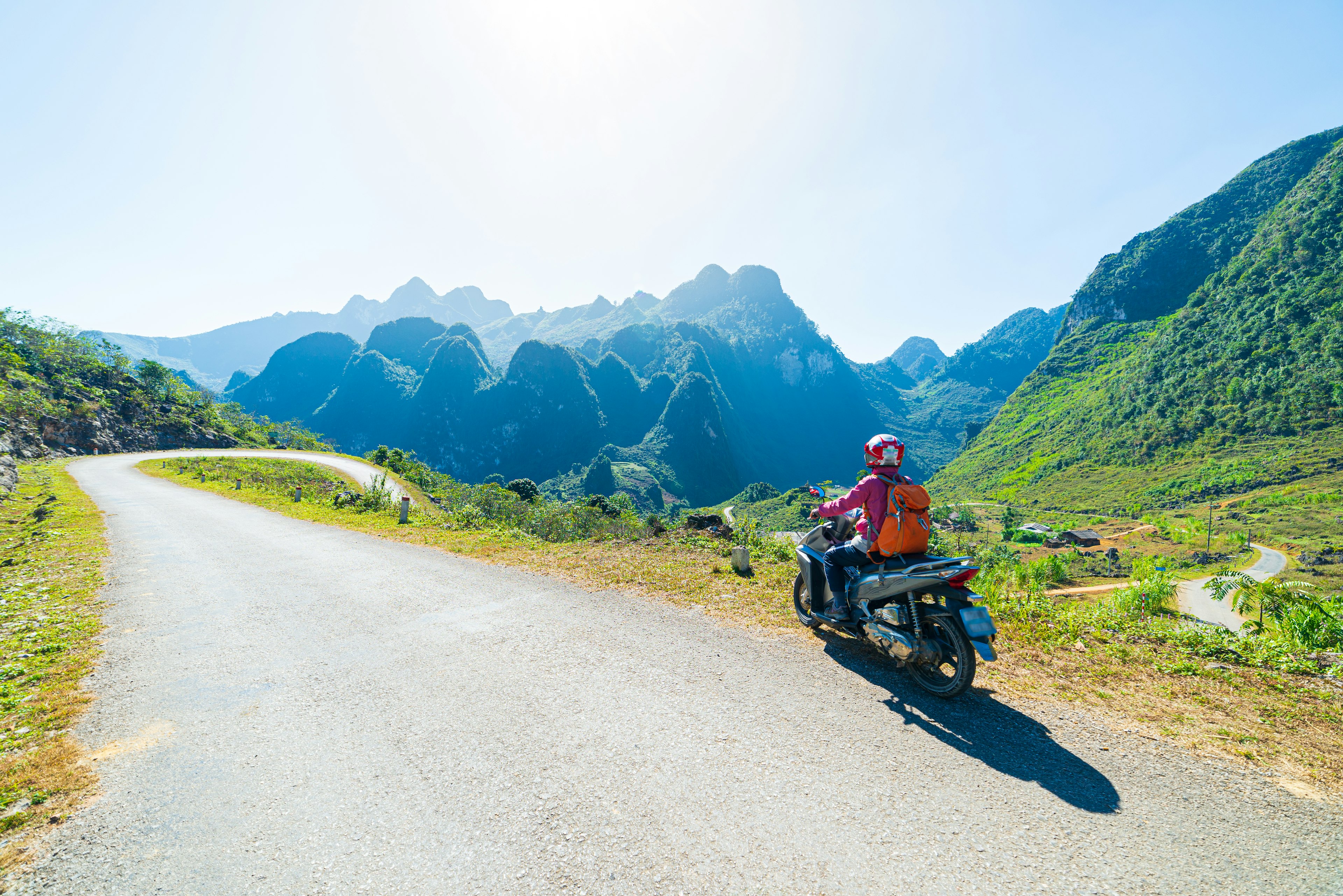 A man on a motor scooter rides on windy mountain road. Hazy green hills are visible in the distance.