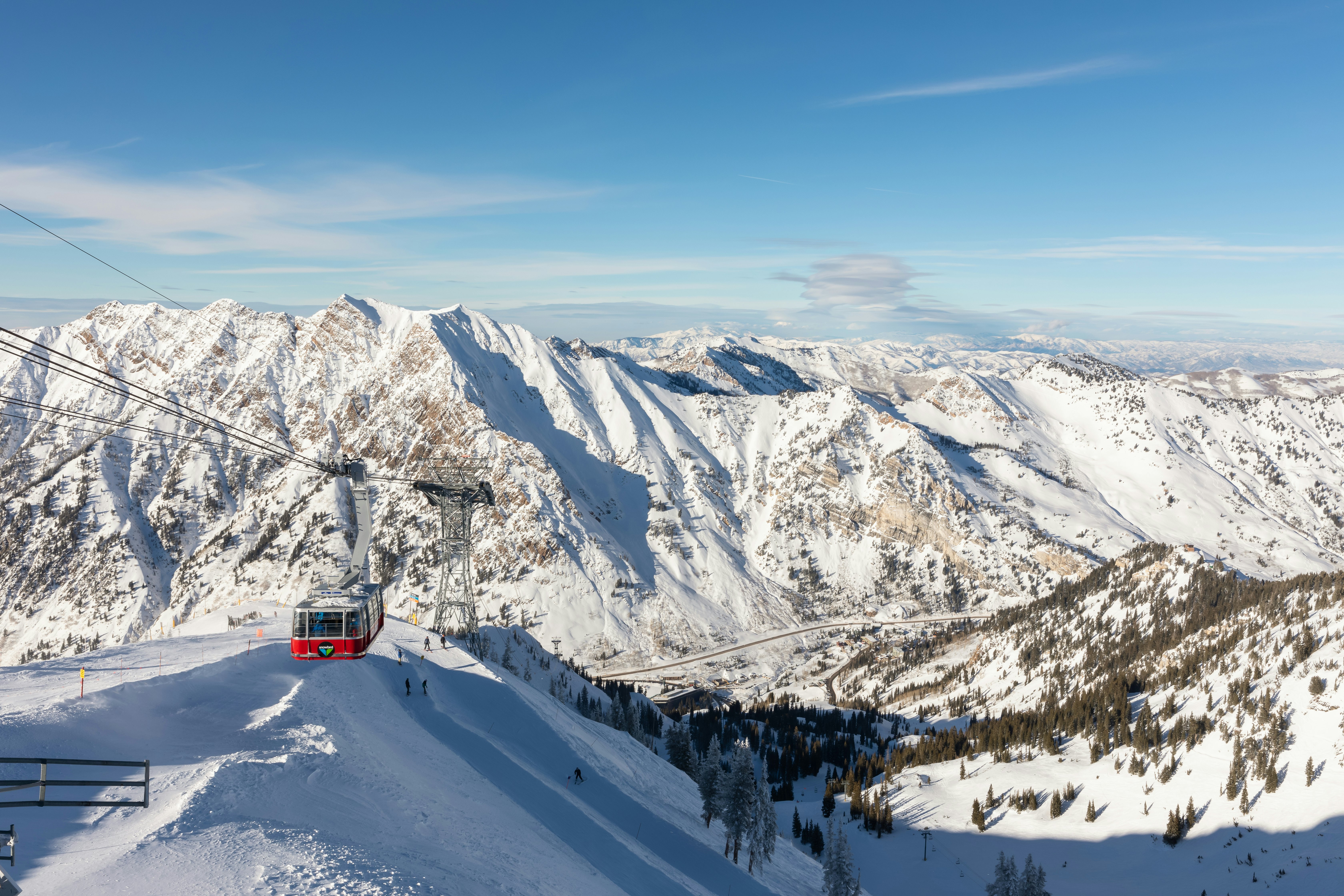 A wide view of an aerial tram ascending the snowy slope of a mountain, with other mountains in the distance.