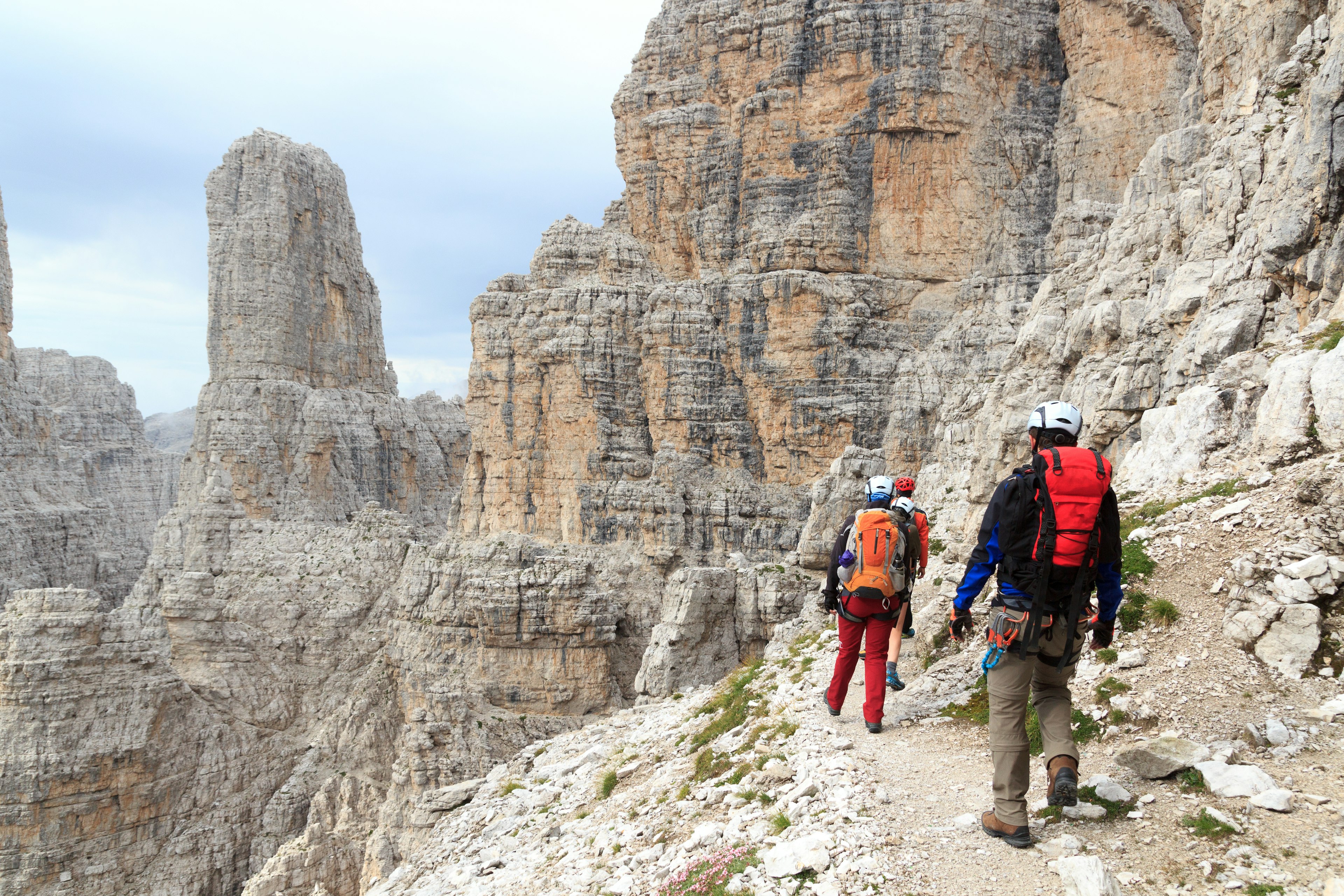 People in helmets walking along a path by a steep cliff in the mountains, with weathered rocky peaks and rock faces surrounding them