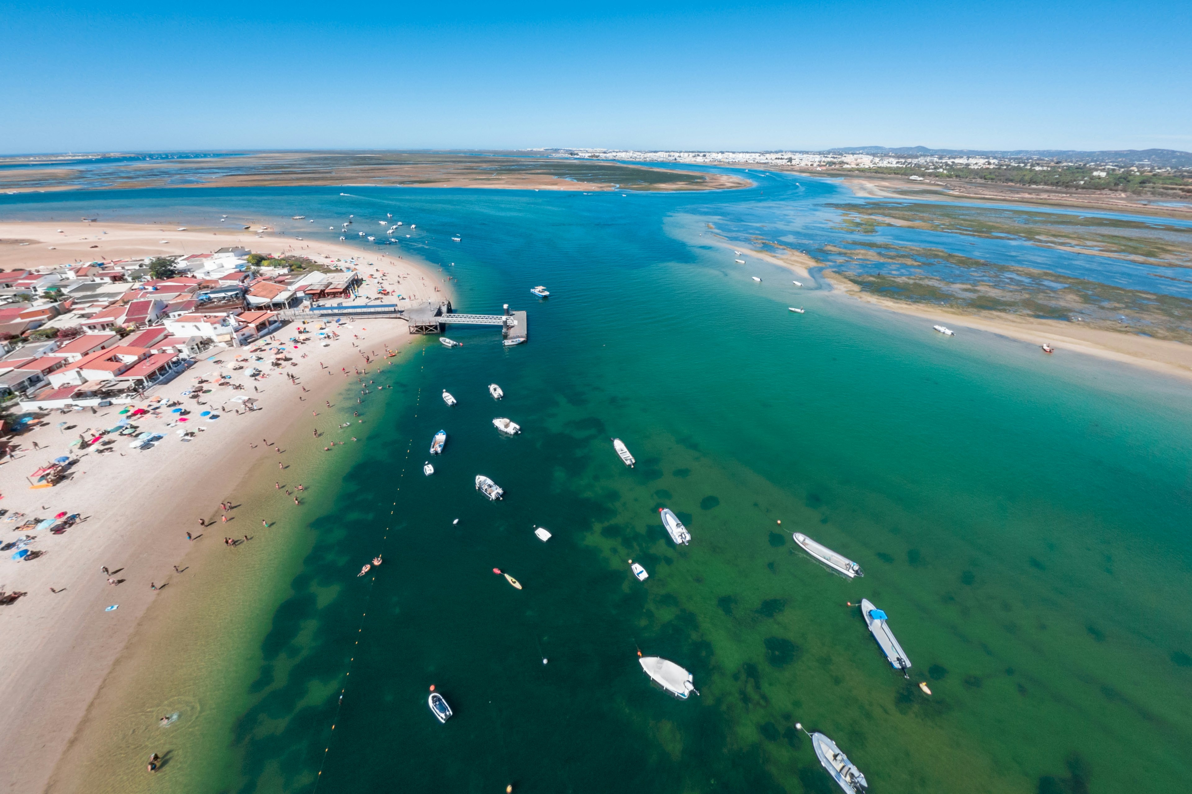 An aerial view of boats moored in the water near Armona Island, with people on a beach by buildings and a pier, Ria Formosa, the Algarve, Portugal