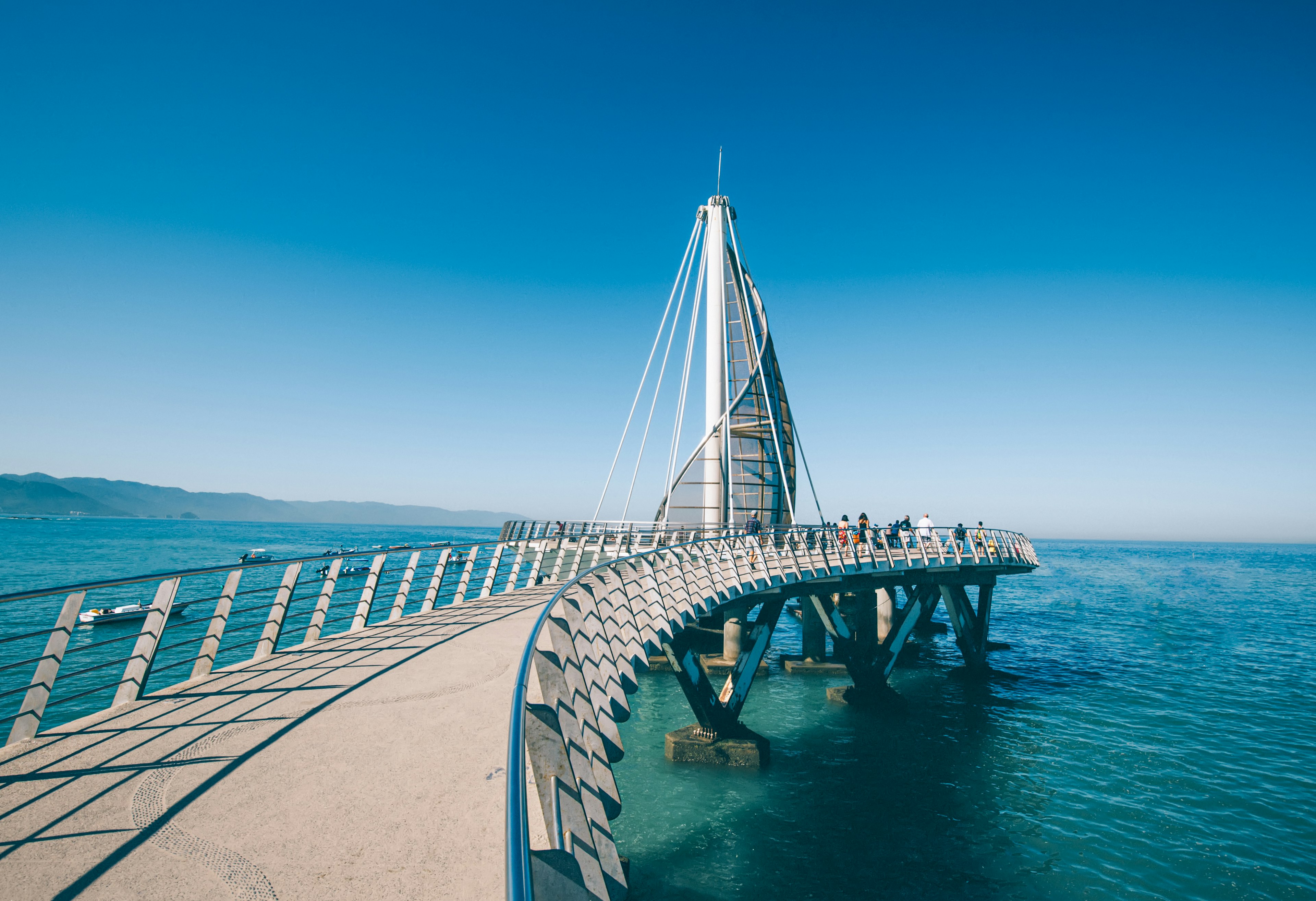 People walk to the end of a a pier built in a modern architectural style that juts into the blue waters of a warm bay