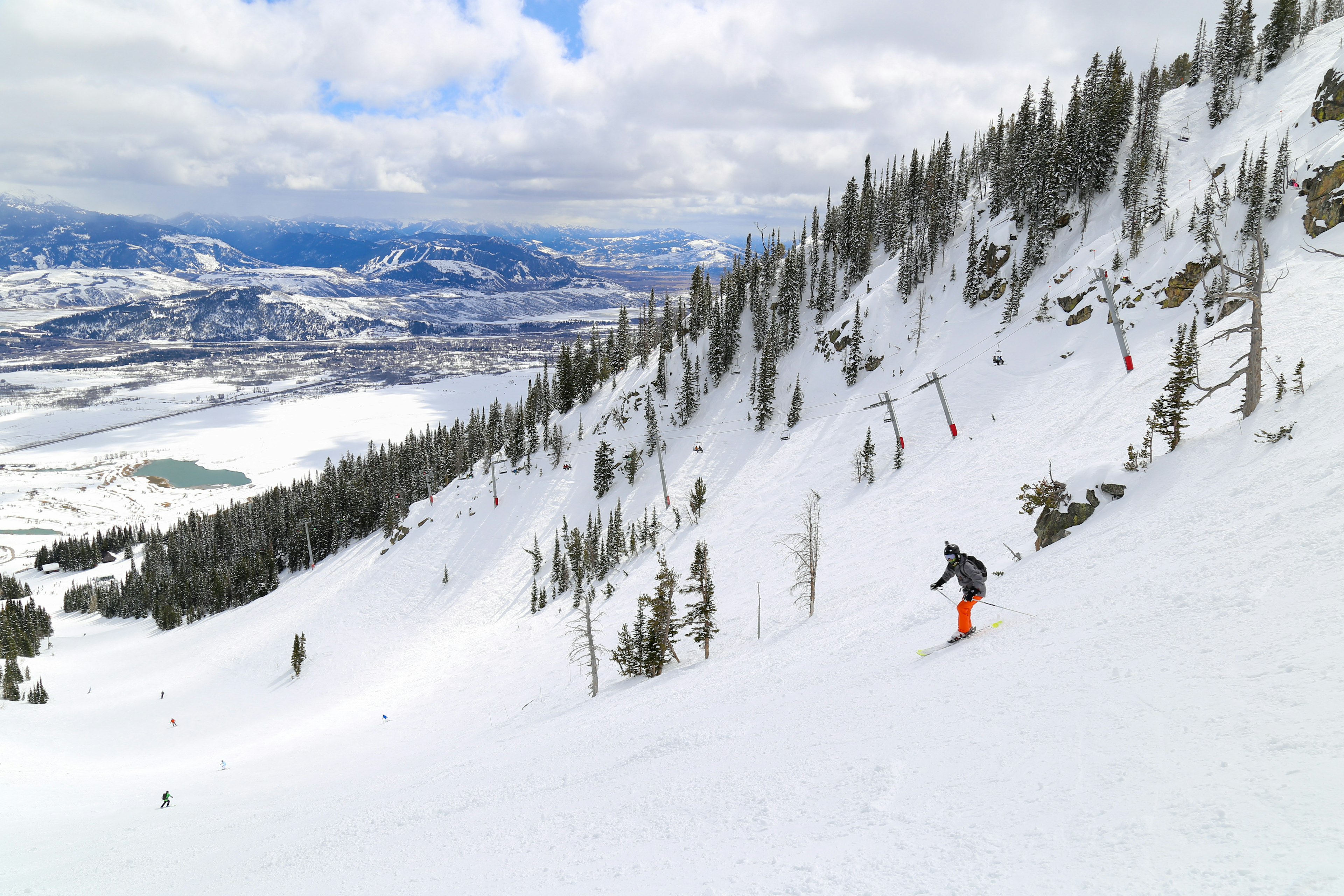 A skier in orange pants skis down the edge of a tree-covered mountain with a mountain range in the background.