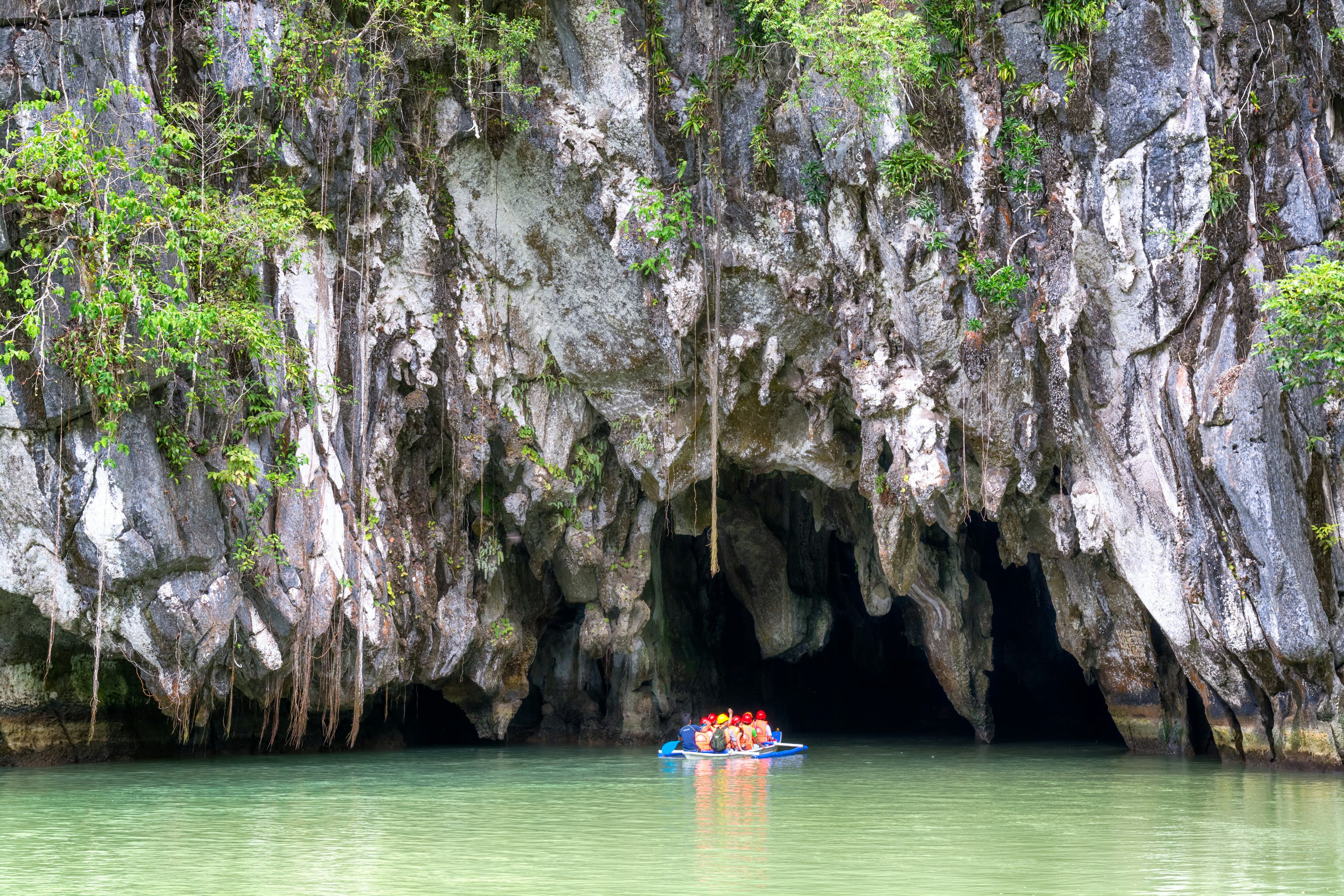 People in a raft enter the entrance to a cavern, which is at the bottom of a rocky cliff covered with vines