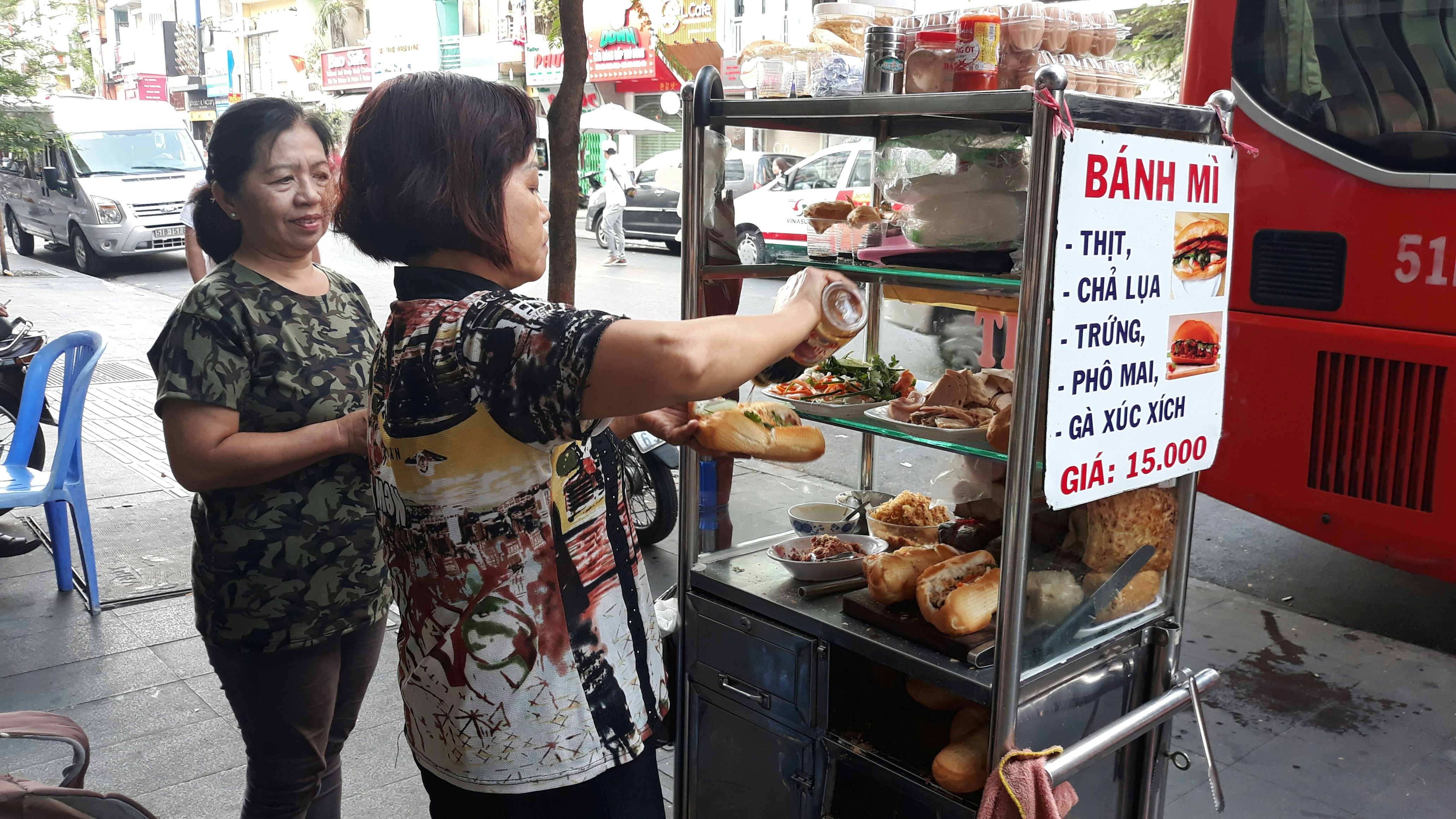 A woman buys a banh mi sandwich from a sidewalk vendor in a busy street in Ho Chi Minh City, Vietnam