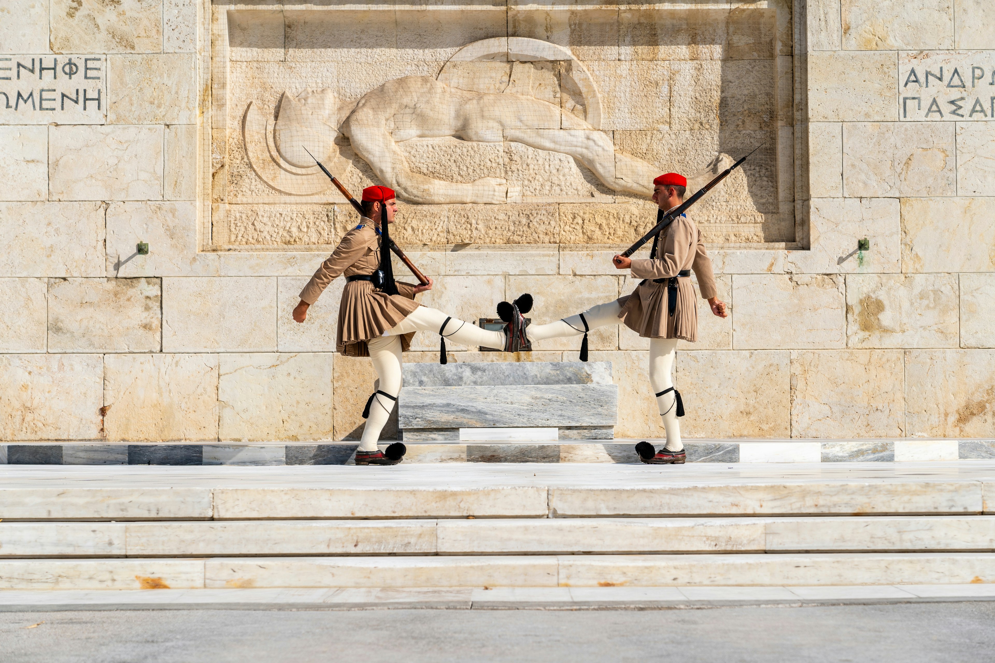 Two soliders in long tunics, white leggings and shoes with pompoms kick their legs high as they march